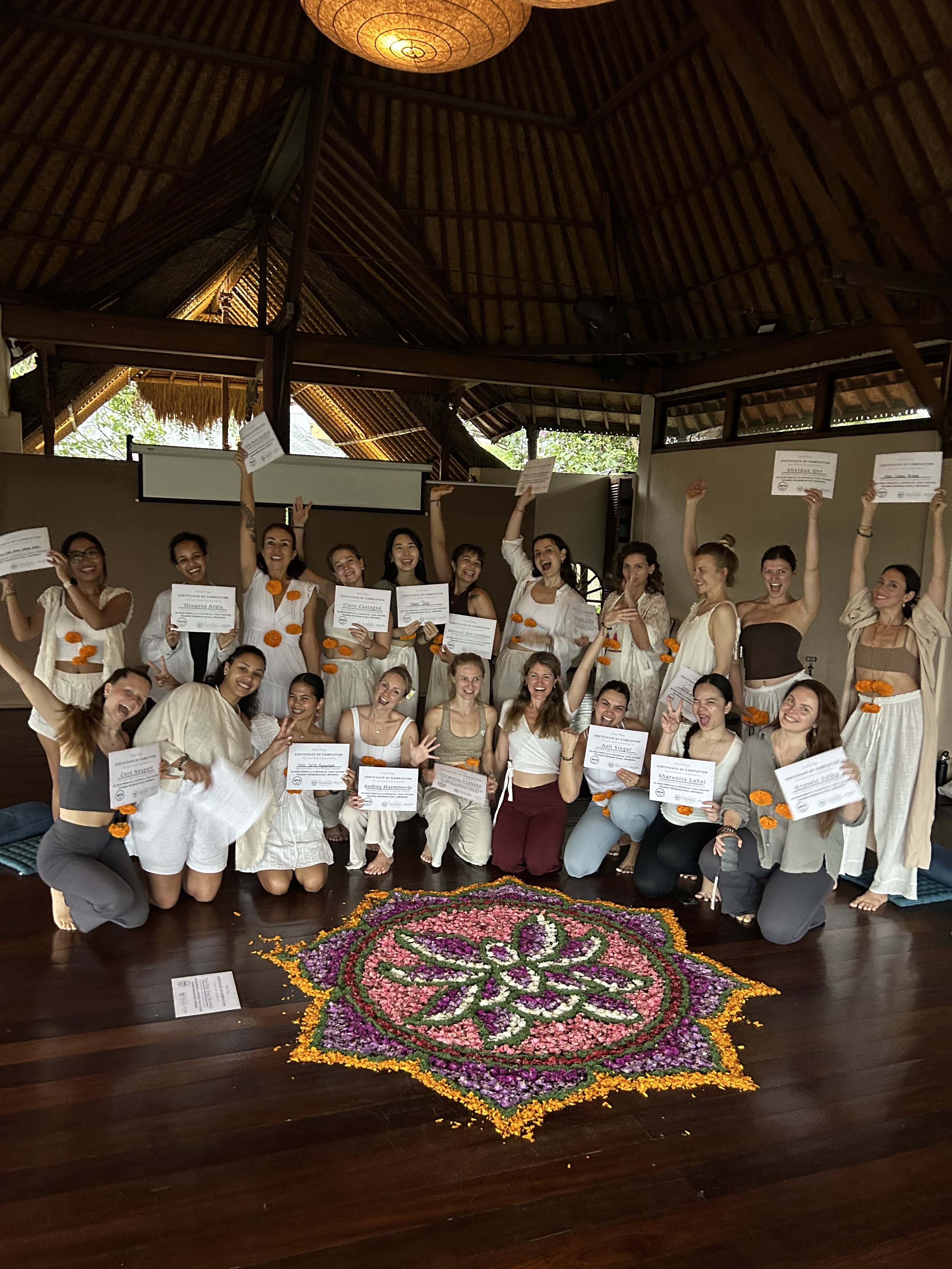 Prenatal Yoga Teacher training students holding certificates around a large, colorful floral design on the floor, in a tropical-style open-air hall with a thatched roof.