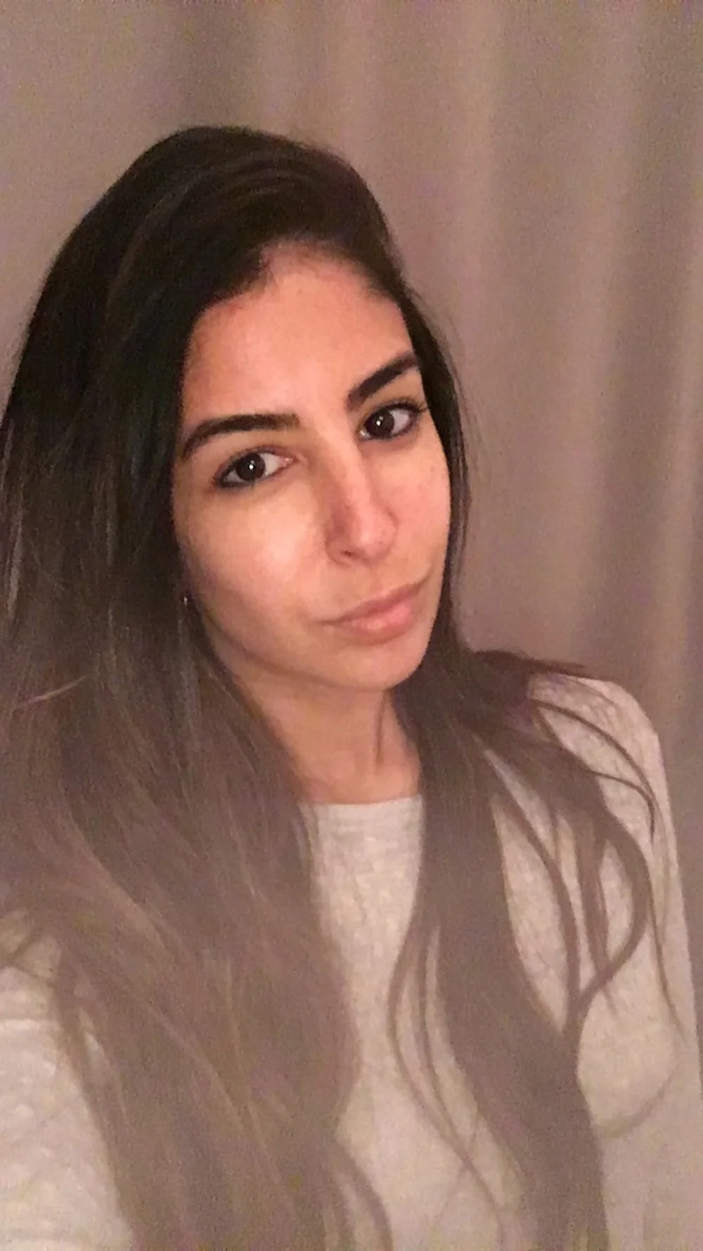 Online prenatal yoga training review. A young woman with long dark hair and light skin taking a selfie indoors, with a neutral-colored wall background.