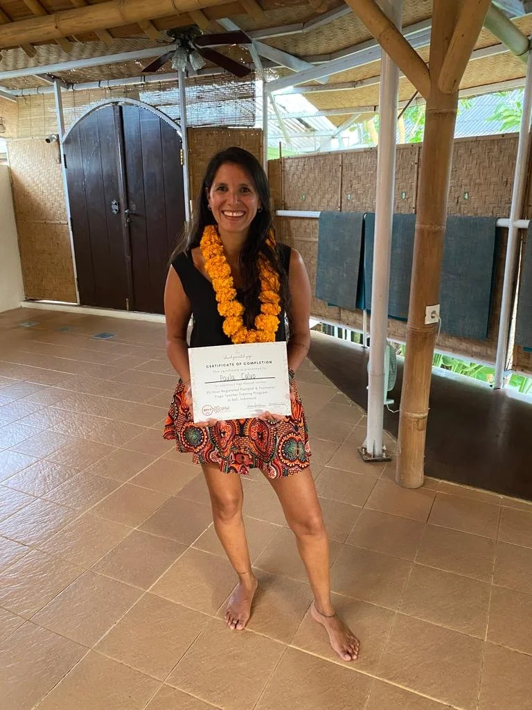 A woman wearing a black sleeveless top and a colorful skirt standing barefoot indoors, smiling, with a marigold flower lei around her neck. She is holding a certificate of completion. The background includes bamboo walls, a thatched roof, and towels hanging on a line.