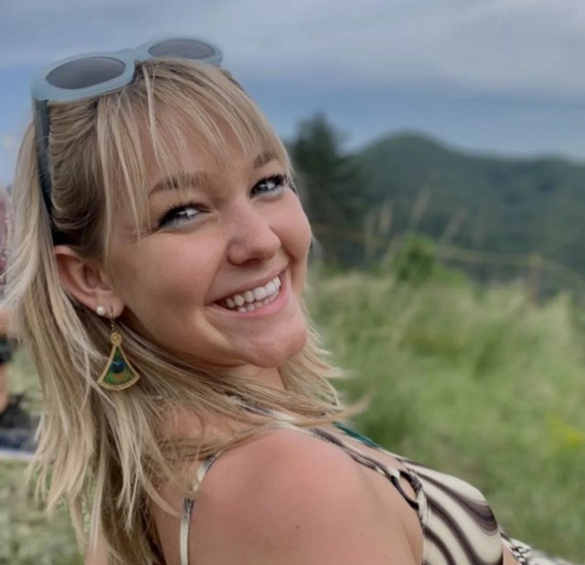 Online prenatal yoga training review. A young woman with blonde hair smiling sideways at the camera outdoors, wearing sunglasses on her head and loose earrings, with mountains and greenery in the background.