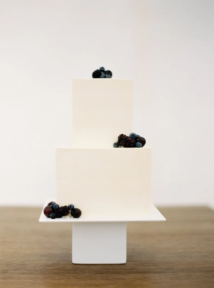Minimalist two-tier white cake with blueberries and blackberries on a square pedestal stand.
