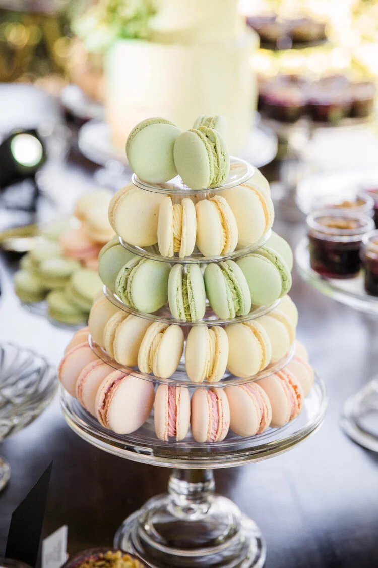 A tiered stand displaying assorted macarons in pastel colors, including green, yellow, and pink, on a glass table.