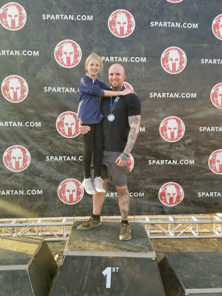 A man with tattoos standing on a first-place podium, holding a young girl who is wearing a medal around her neck, in front of a sports-themed backdrop with Spartan branding.