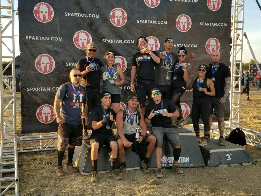 A group of people in athletic gear and medals gathered on a podium after a Spartan race, with a backdrop displaying the Spartan logo and website.