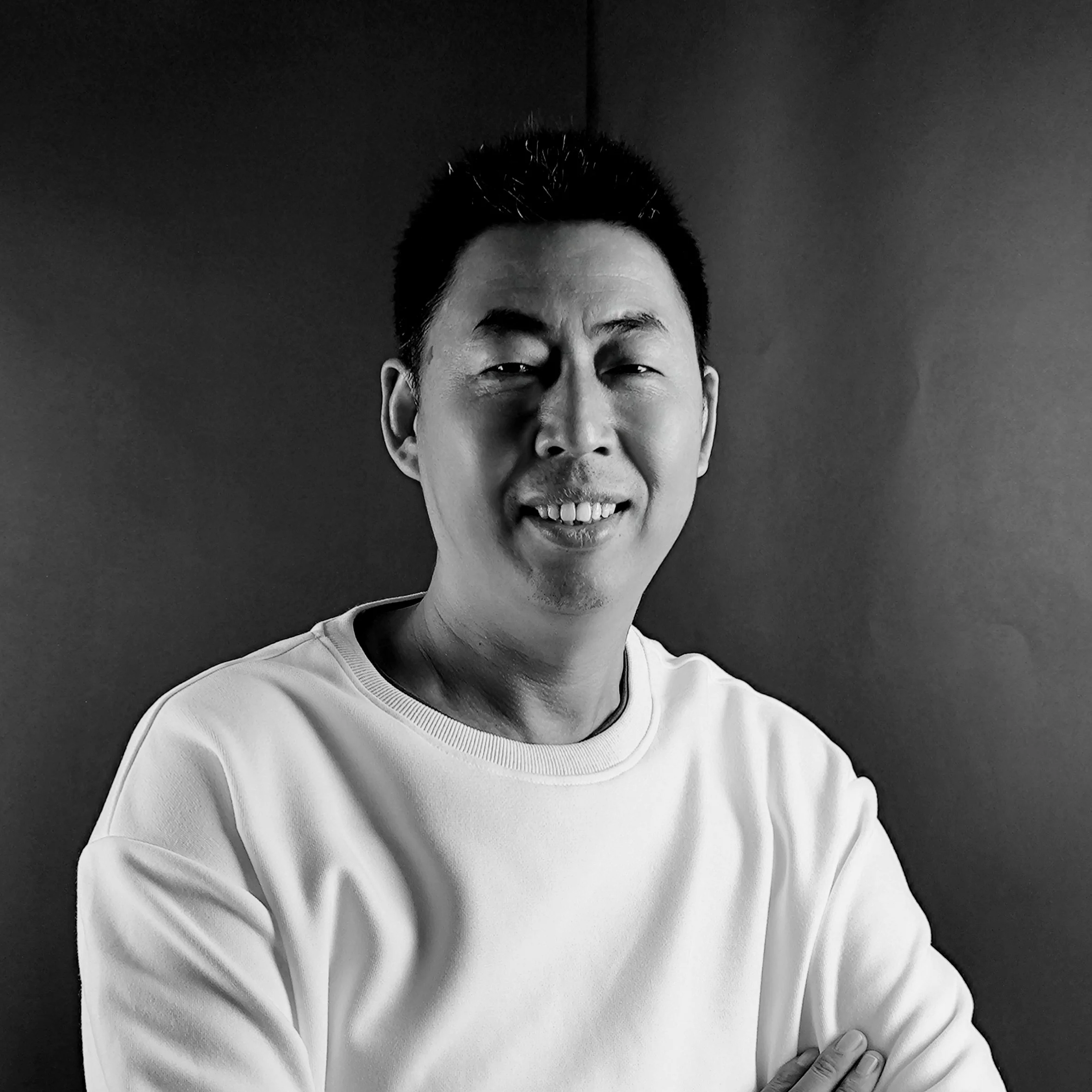 Black and white portrait of a smiling man with short dark hair, wearing a white sweatshirt, standing against a dark background.