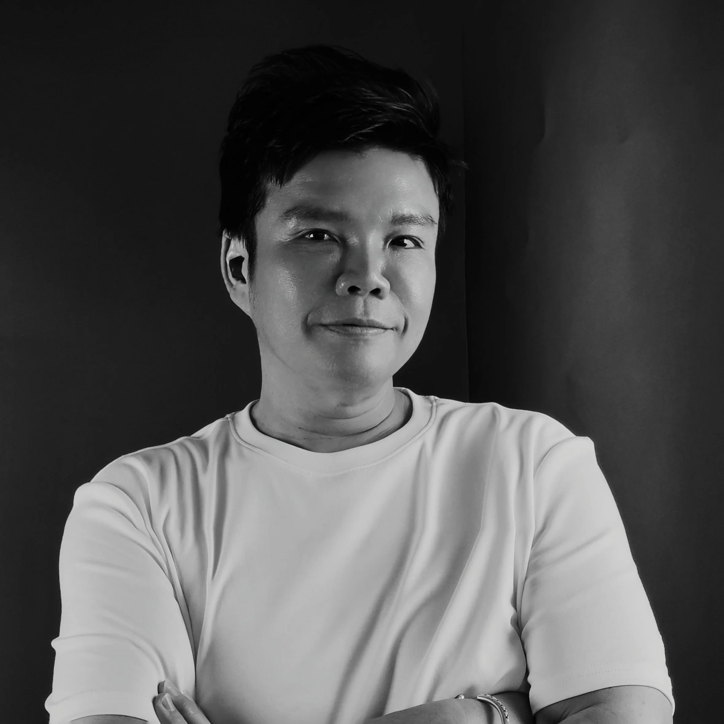 Black and white portrait of a young man with dark hair, wearing a white T-shirt, smiling with arms crossed, against a plain dark background.