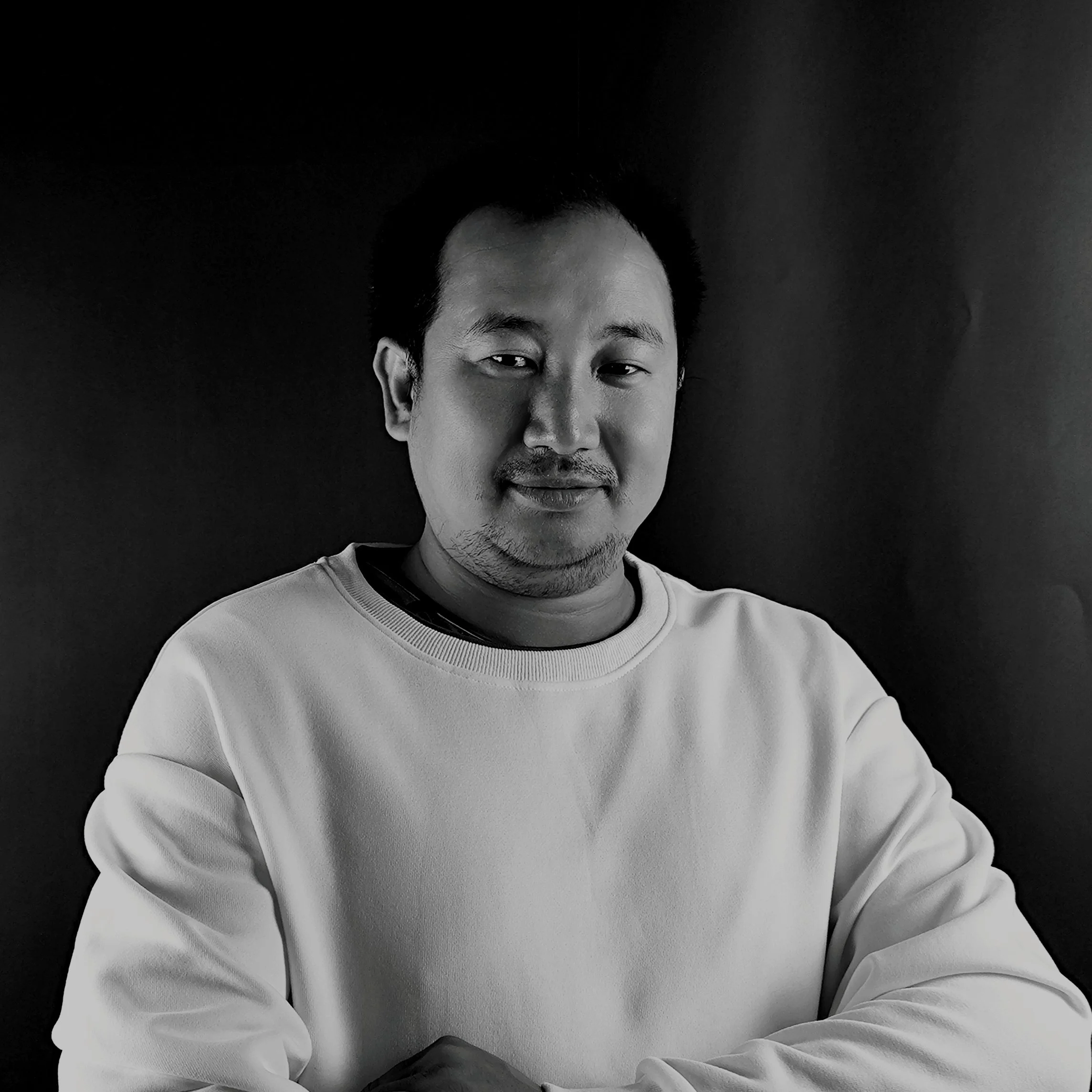 Black and white portrait of a man smiling, wearing a light-colored sweatshirt, with a dark background.