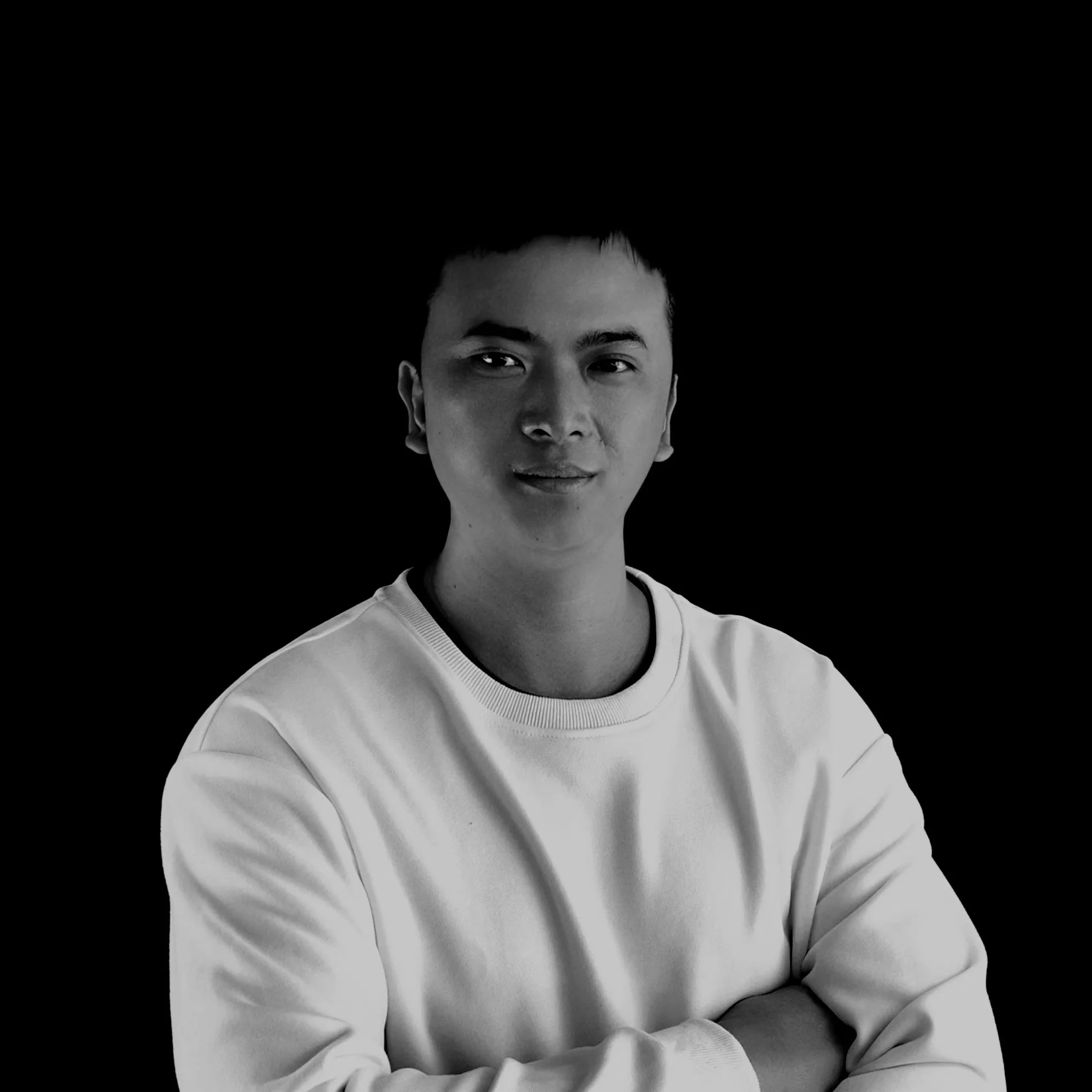 Black-and-white portrait of a young man with short hair, wearing a light-colored sweatshirt, smiling with arms crossed against a dark background.