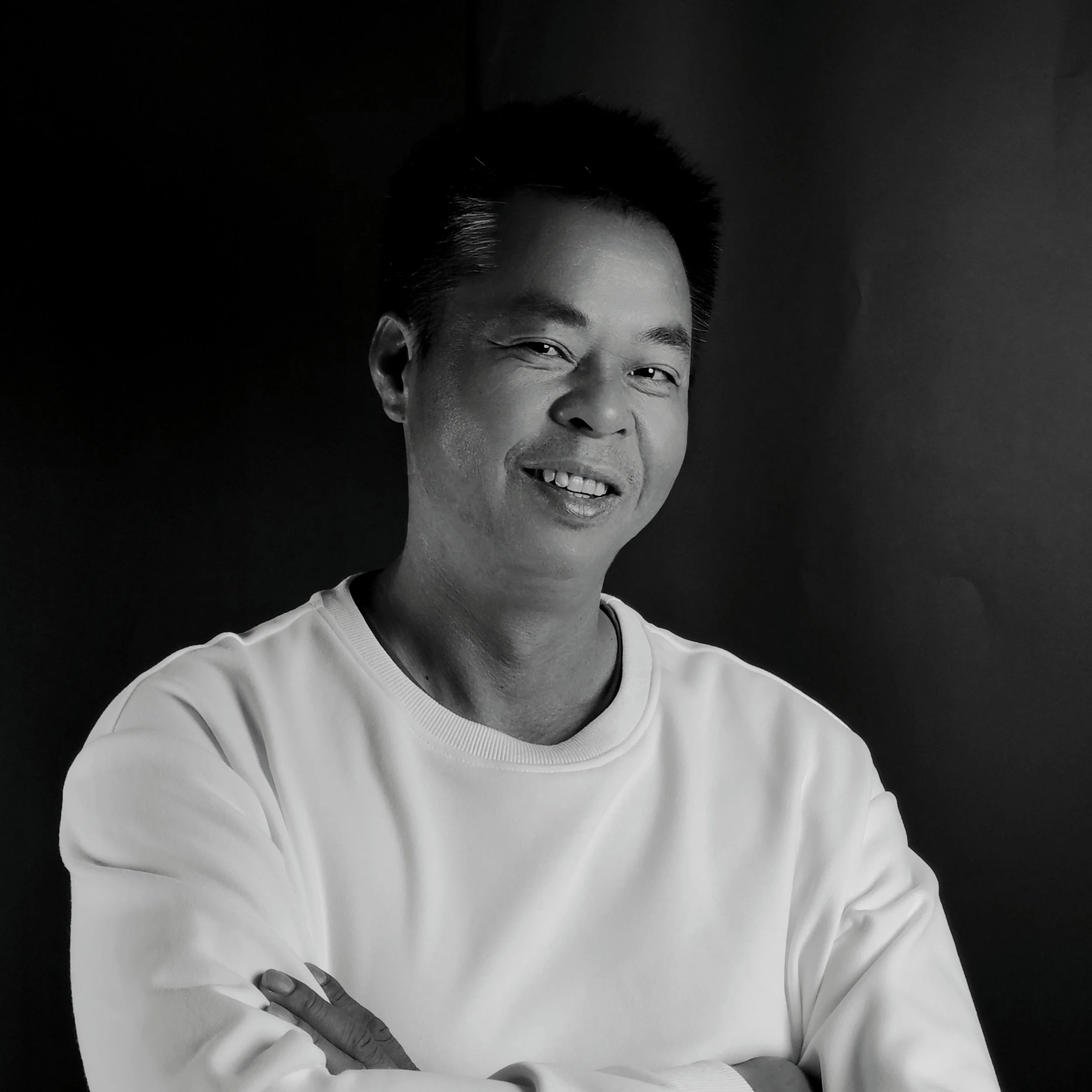 Black and white portrait of a smiling man with arms crossed, wearing a white shirt, against a dark background.
