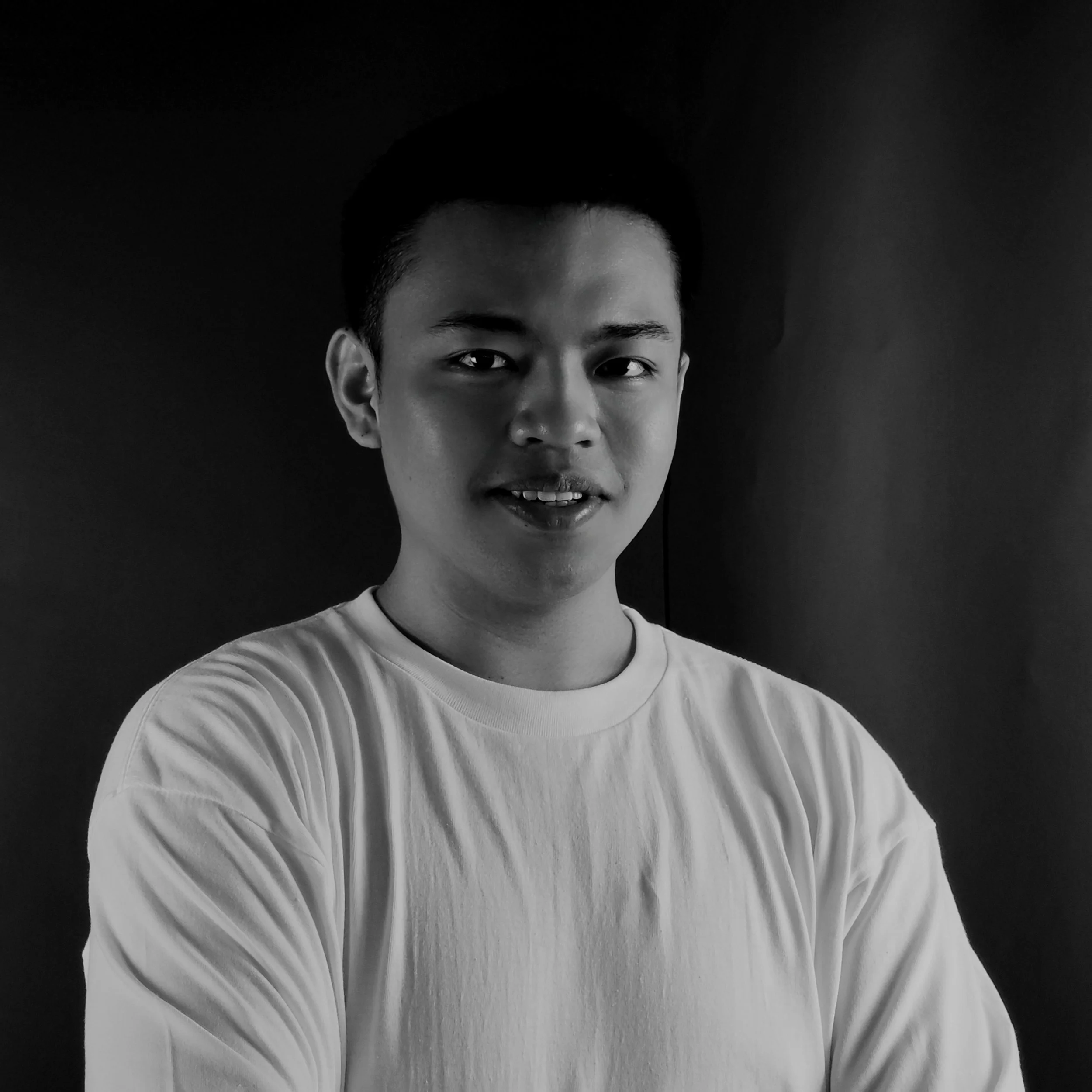 Black and white portrait of a young man with short dark hair, wearing a light-colored t-shirt, facing the camera with a slight smile against a dark background.