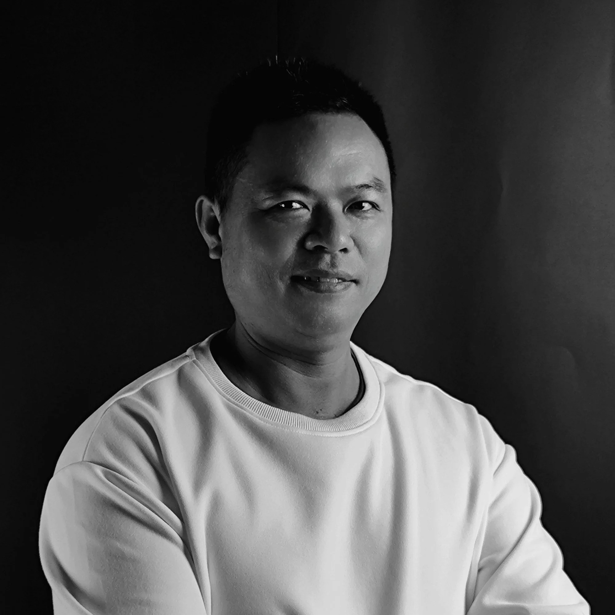 Black and white portrait of a man with short dark hair, wearing a light-colored sweatshirt, smiling slightly, against a plain dark background.