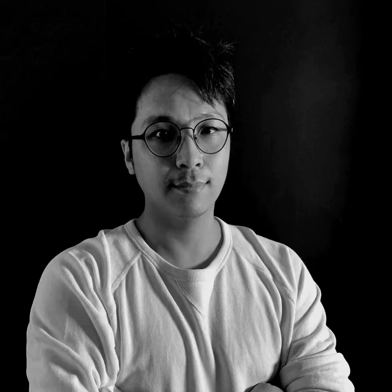Black and white portrait of a young man with glasses, wearing a light-colored sweatshirt, against a dark background.