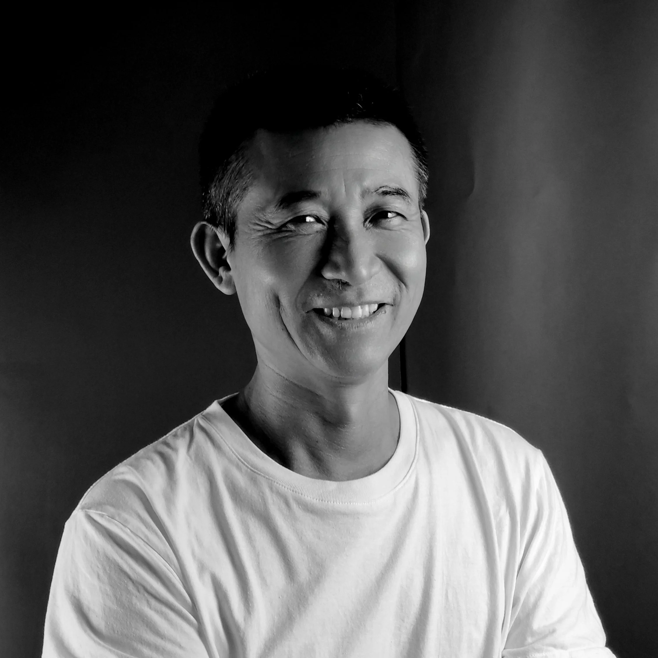 Black and white portrait of a smiling man in a white shirt, with a playful expression and one eye squinting.