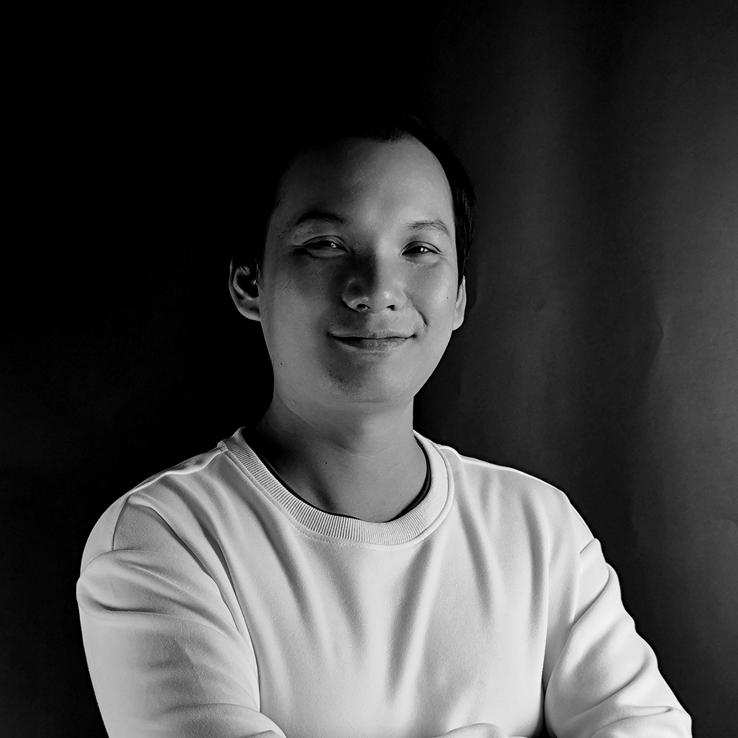 Black and white photo of a young man with short hair, smiling, wearing a light-colored sweatshirt, and posing against a dark background.
