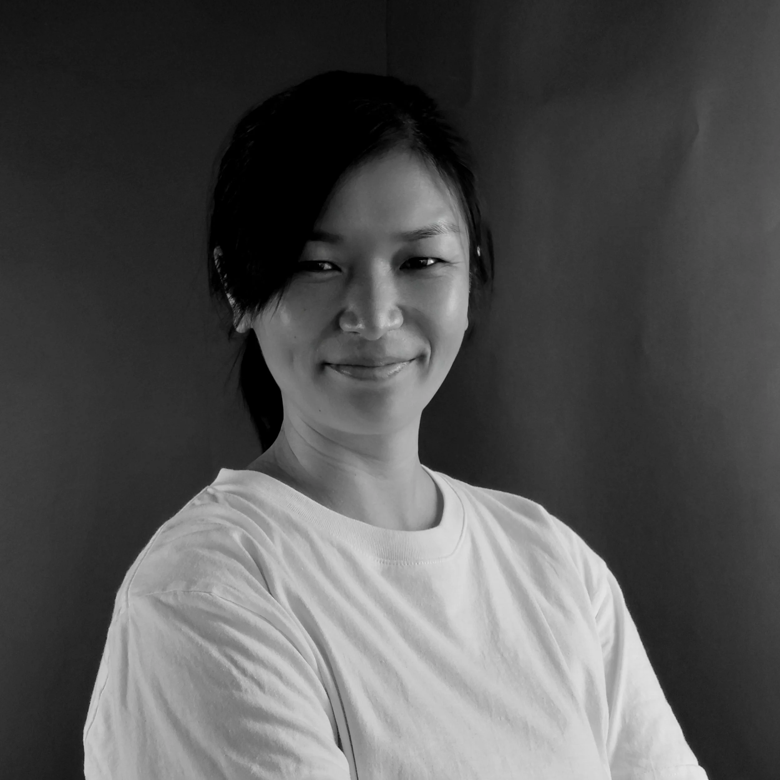 A woman with short hair smiling and looking at the camera, wearing a white T-shirt, photographed in black and white.