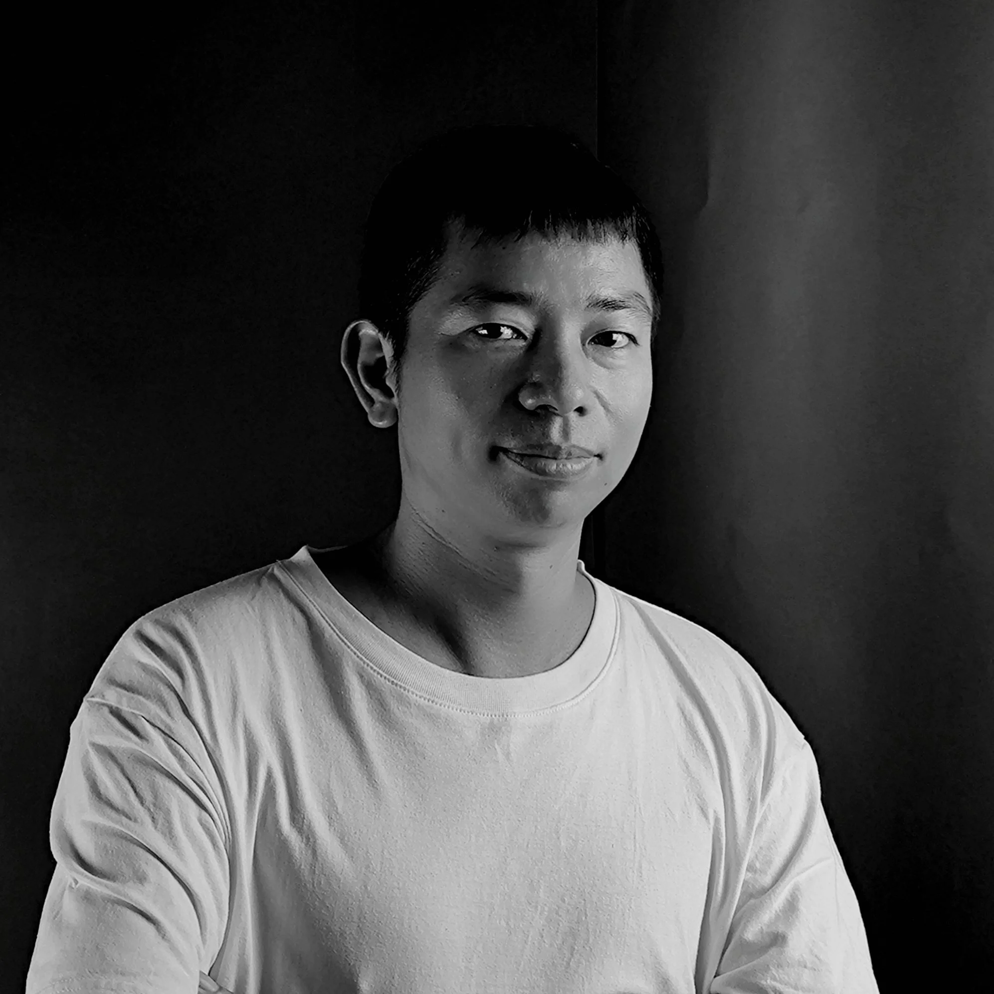 Black and white portrait of a young man with short hair, wearing a white shirt, smiling slightly, standing against a dark background.