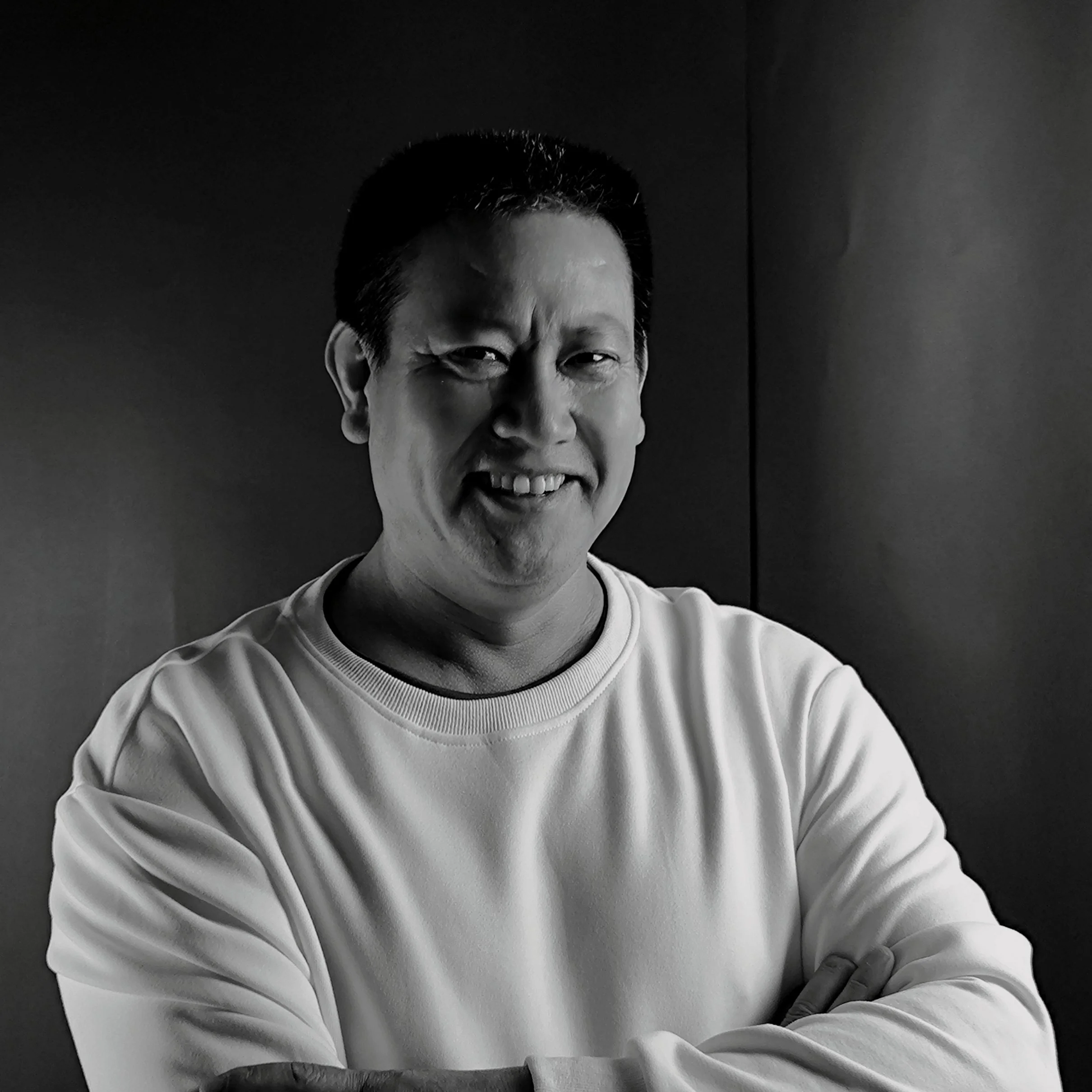 Black and white photo of a man smiling, with arms crossed, wearing a light-colored sweatshirt, standing against a plain background.