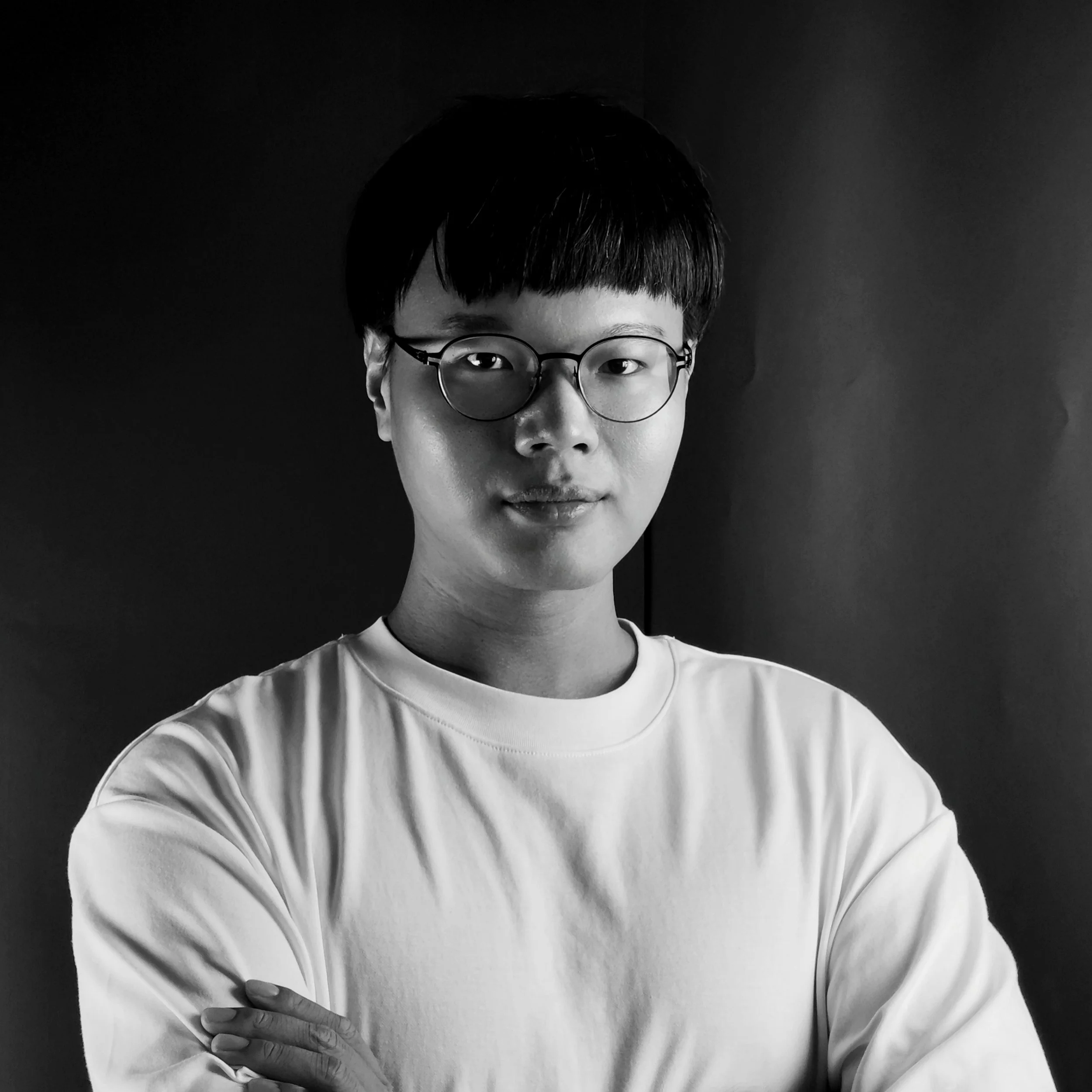 Black and white portrait of a person with short dark hair, wearing glasses and a light-colored t-shirt, crossing their arms and looking at the camera against a dark background.
