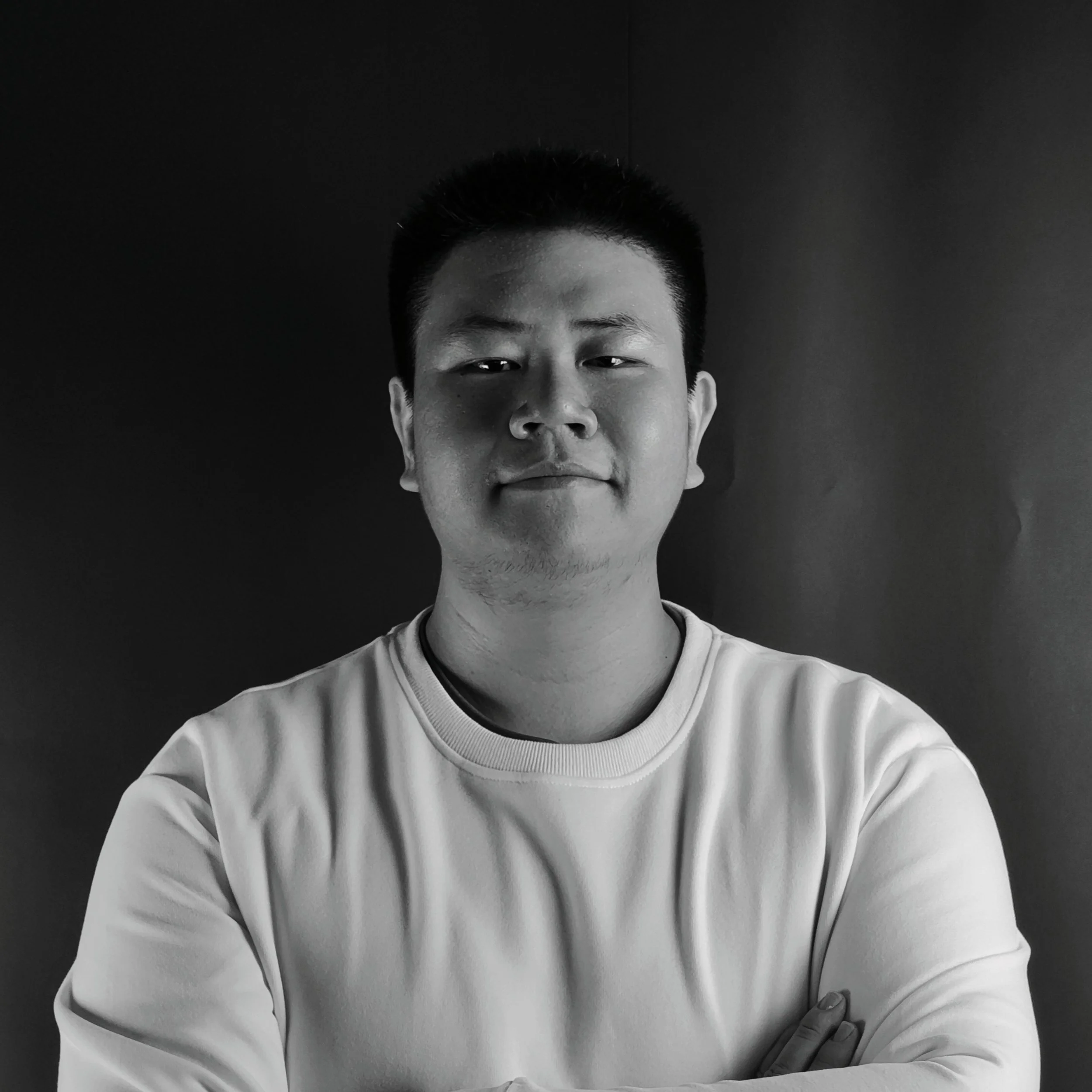 Black and white portrait of a young man with crossed arms, smiling slightly, facing the camera against a dark background.