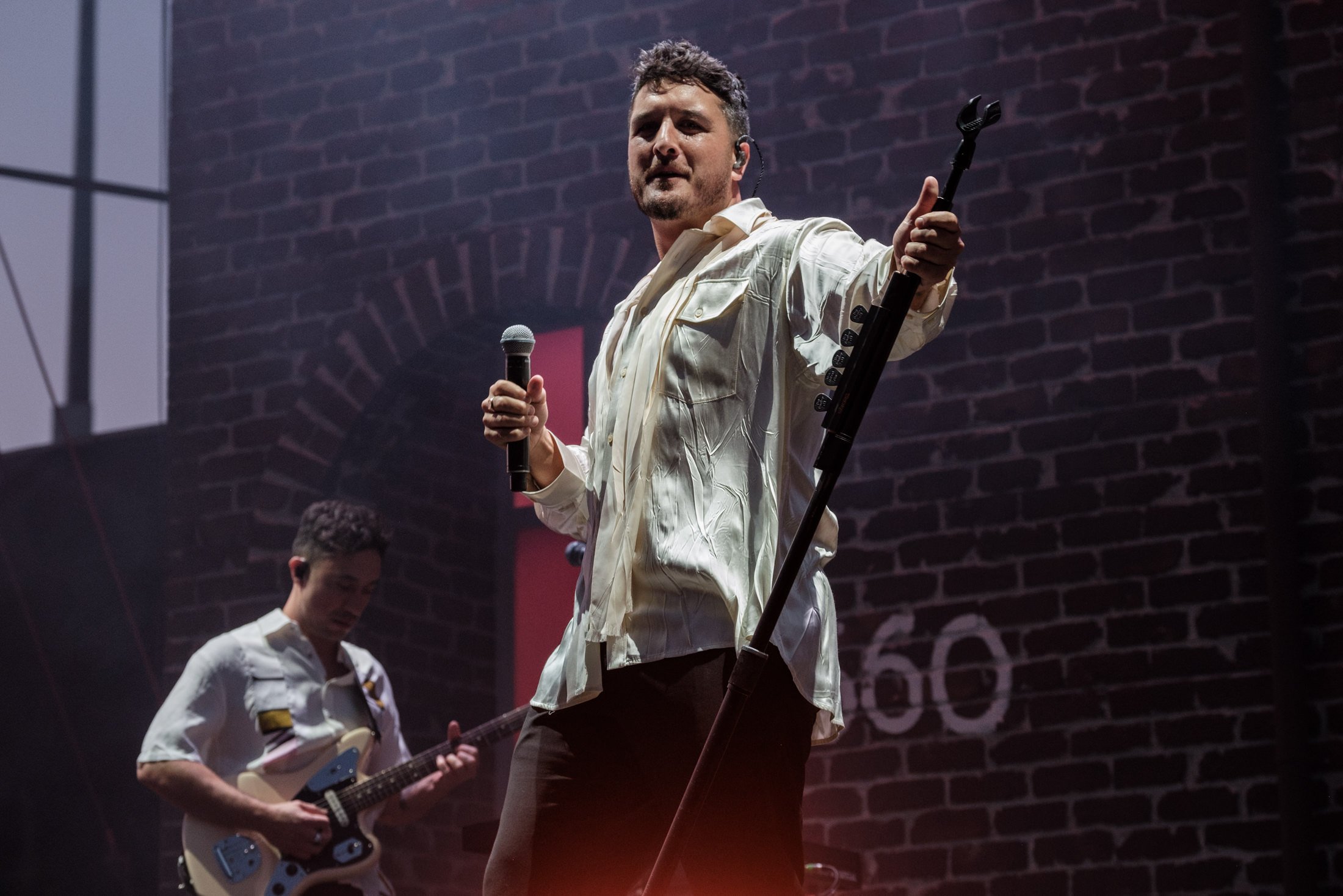 A male singer holding a microphone in his left hand and an electric guitar stand in his right hand, performing on stage with a guitarist in the background against a brick wall.