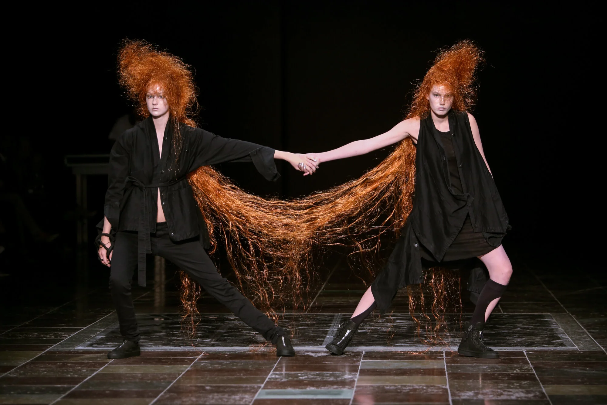 Two models with long, curly red hair holding hands on a runway, wearing black outfits in a fashion show.