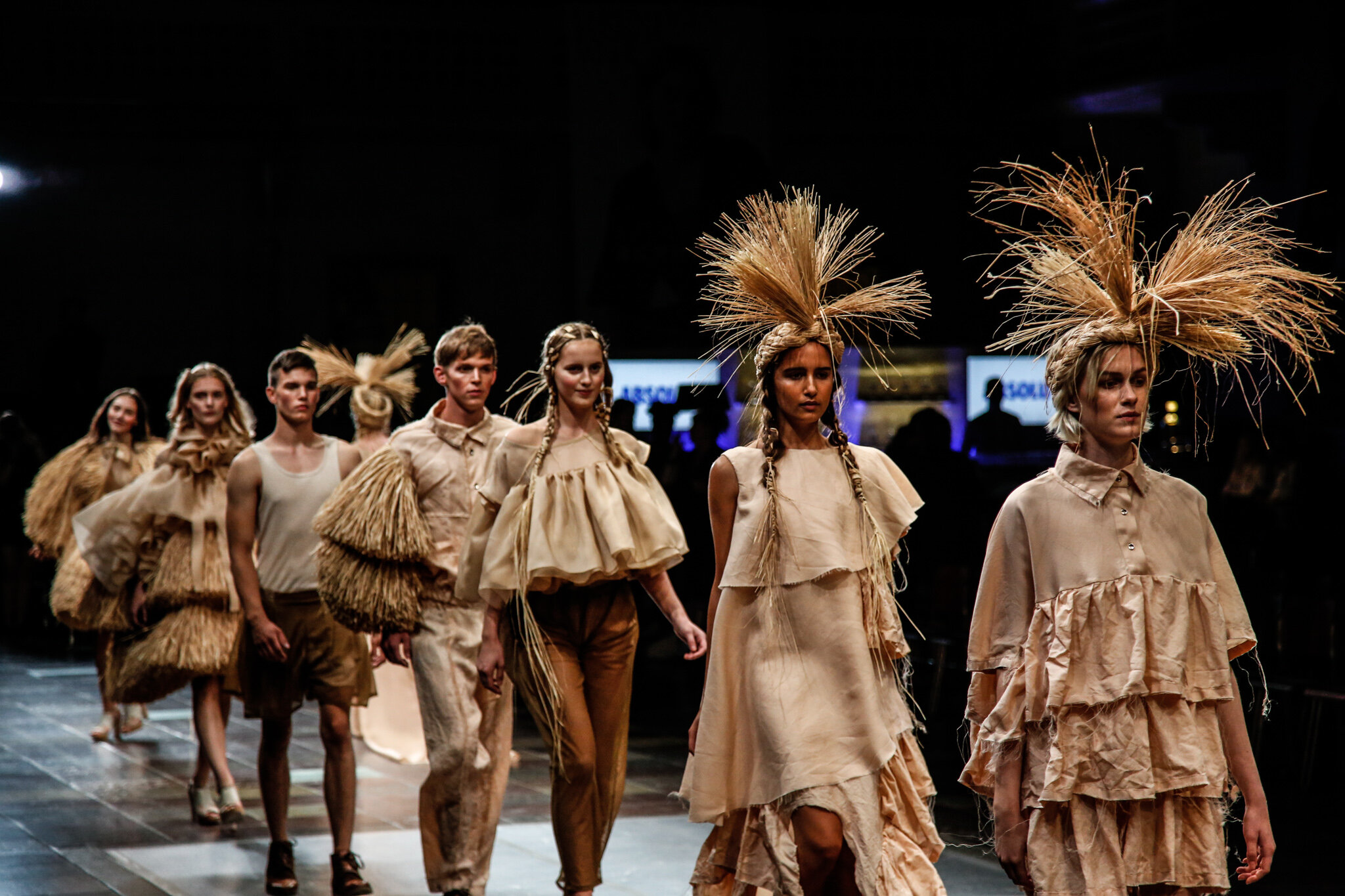 Models walking down a runway during a fashion show, wearing beige and earth-toned clothing with large, spiked accessories on their heads.