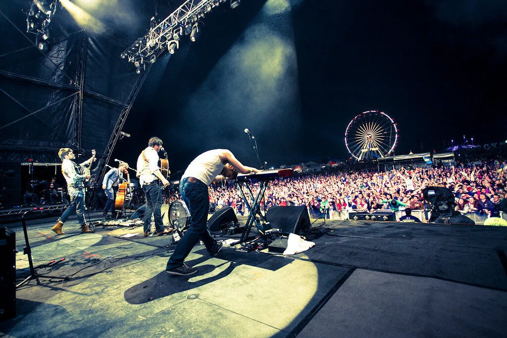A band performing live on stage at a concert with a large audience in front. A ferris wheel is visible in the background at night.