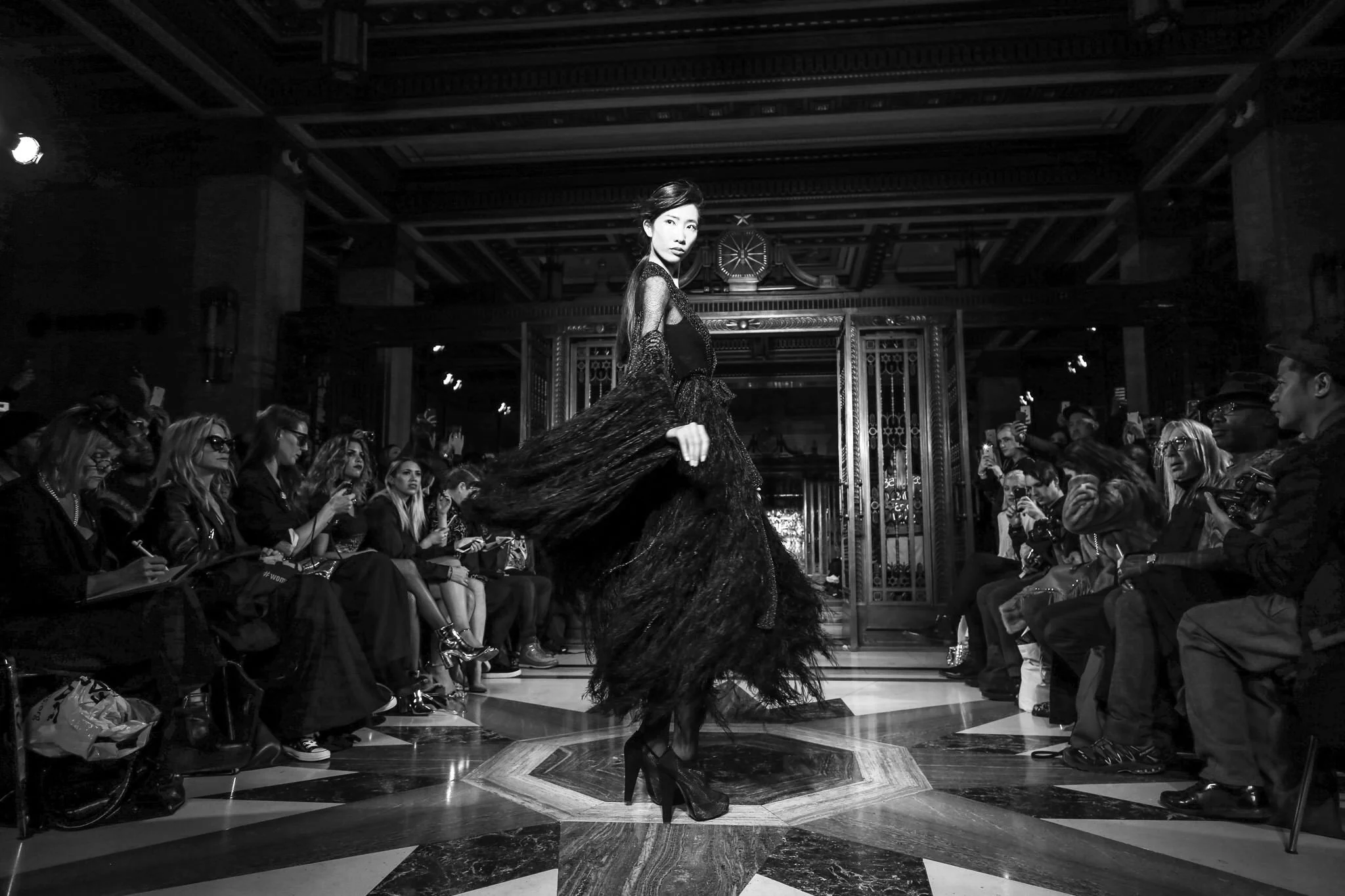 Fashion model walking down runway at a fashion show, surrounded by seated audience members taking photos, in an elegant, ornate interior.