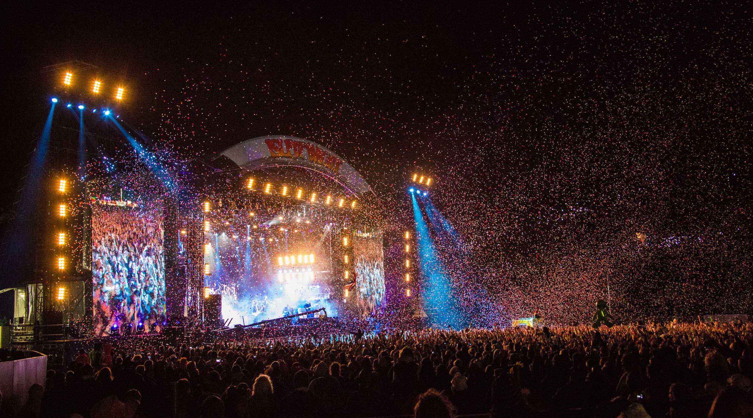 Nighttime outdoor concert with a large crowd, stage lighting, colorful confetti in the air, and two big screens showing the performers.
