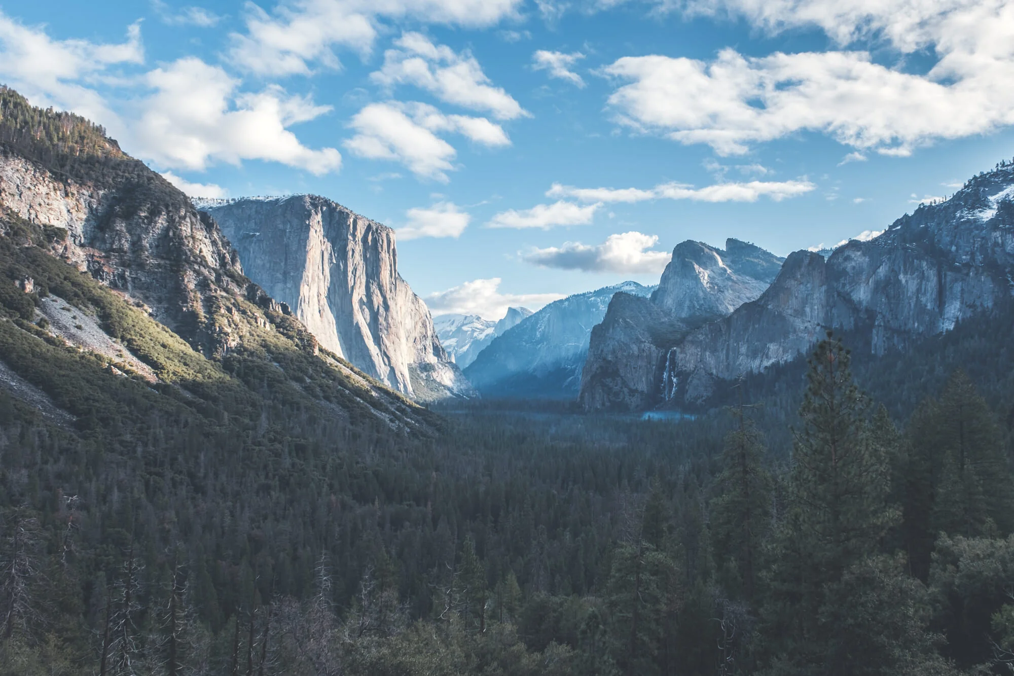 A view of a mountain valley with towering cliffs, green forests, and a sky with clouds.