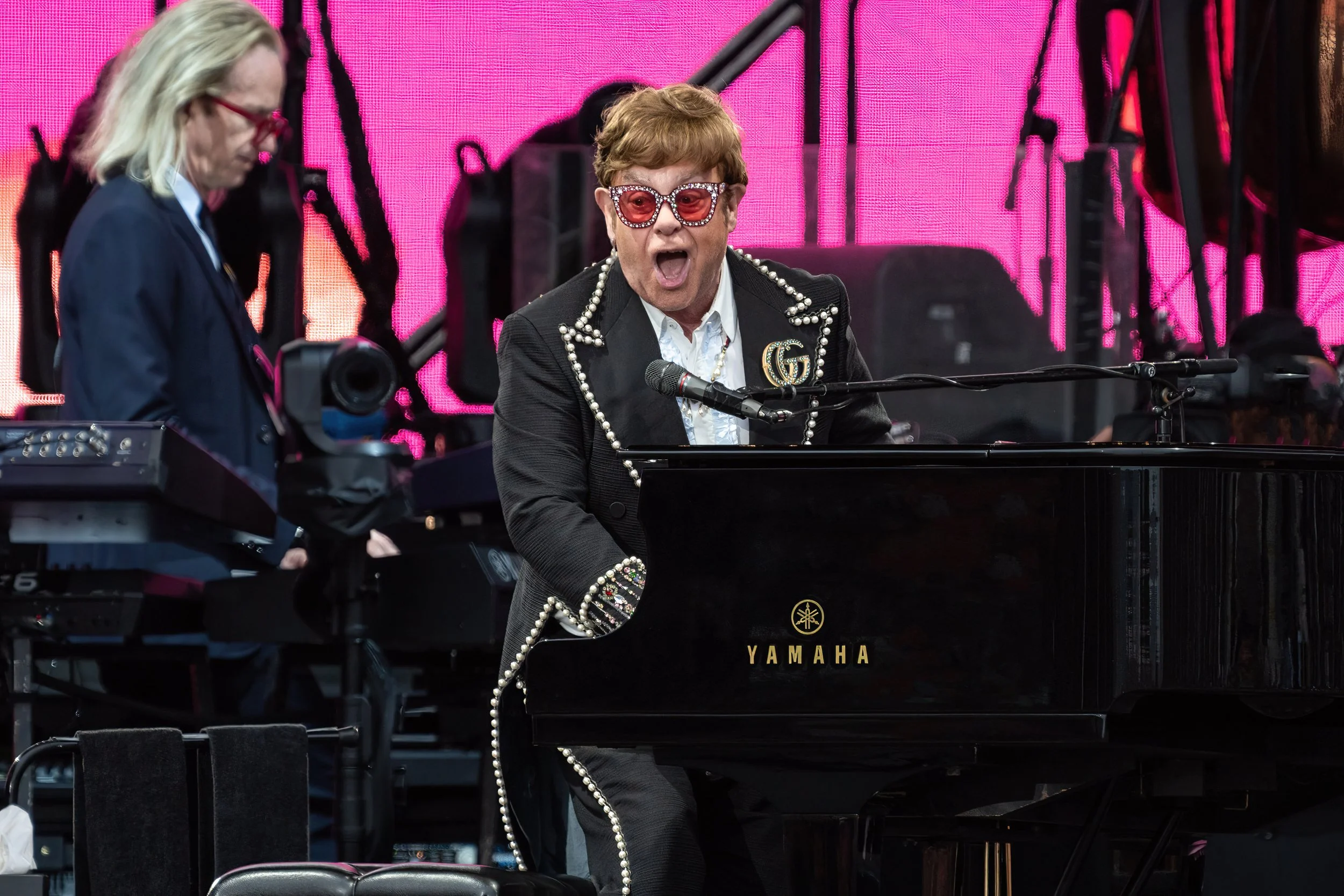 Elton John performing on stage at a concert, wearing a black suit with pearl embellishments, red sunglasses with rhinestones, singing passionately into a microphone while playing a grand Yamaha piano.