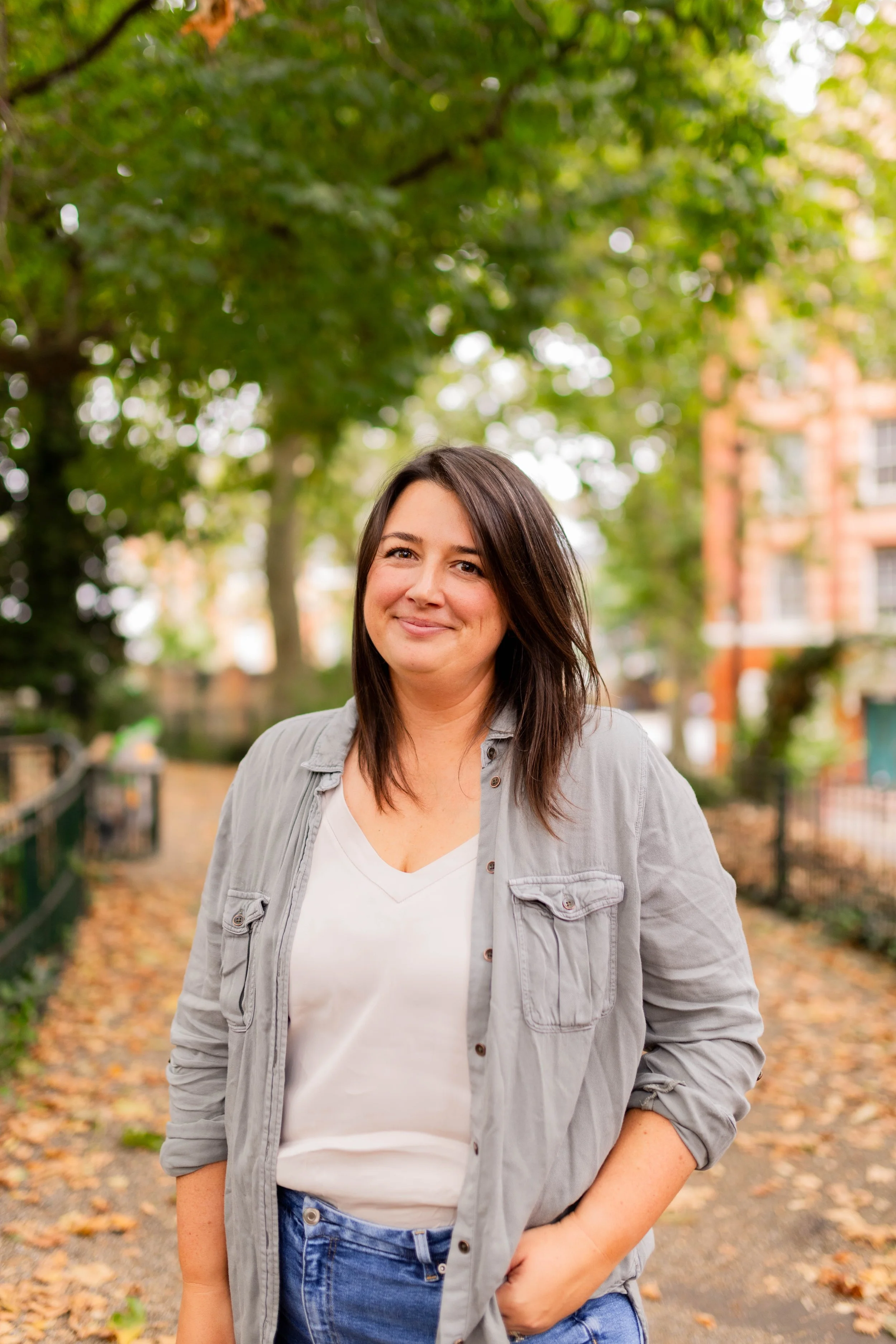 Smiling woman with dark hair in casual gray jacket and white shirt, standing outdoors on a tree-lined pathway with autumn leaves, city buildings in the background.