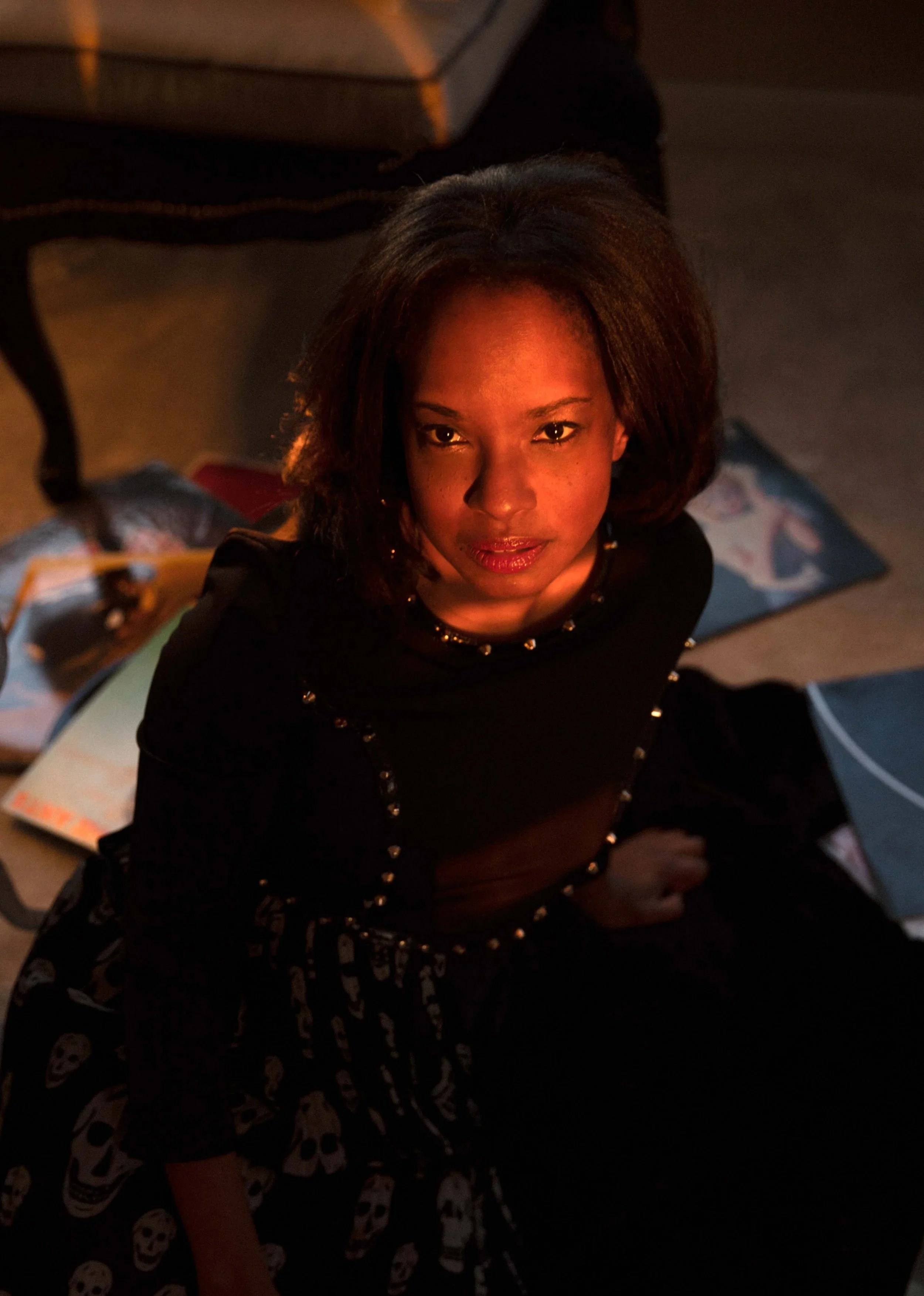 A woman with dark hair and a confident expression looking up at the camera, sitting on the floor surrounded by magazines or books, in a dimly lit room.
