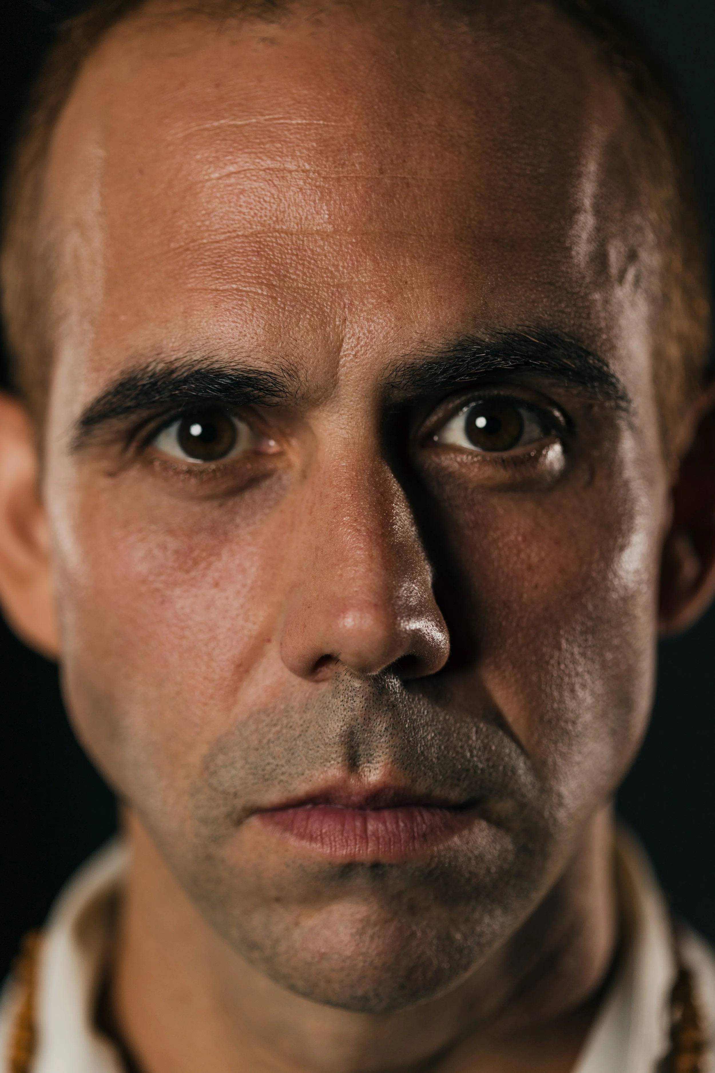 Close-up of a man's face showing intense eyes, dark eyebrows, and detailed skin texture, with a dark background.