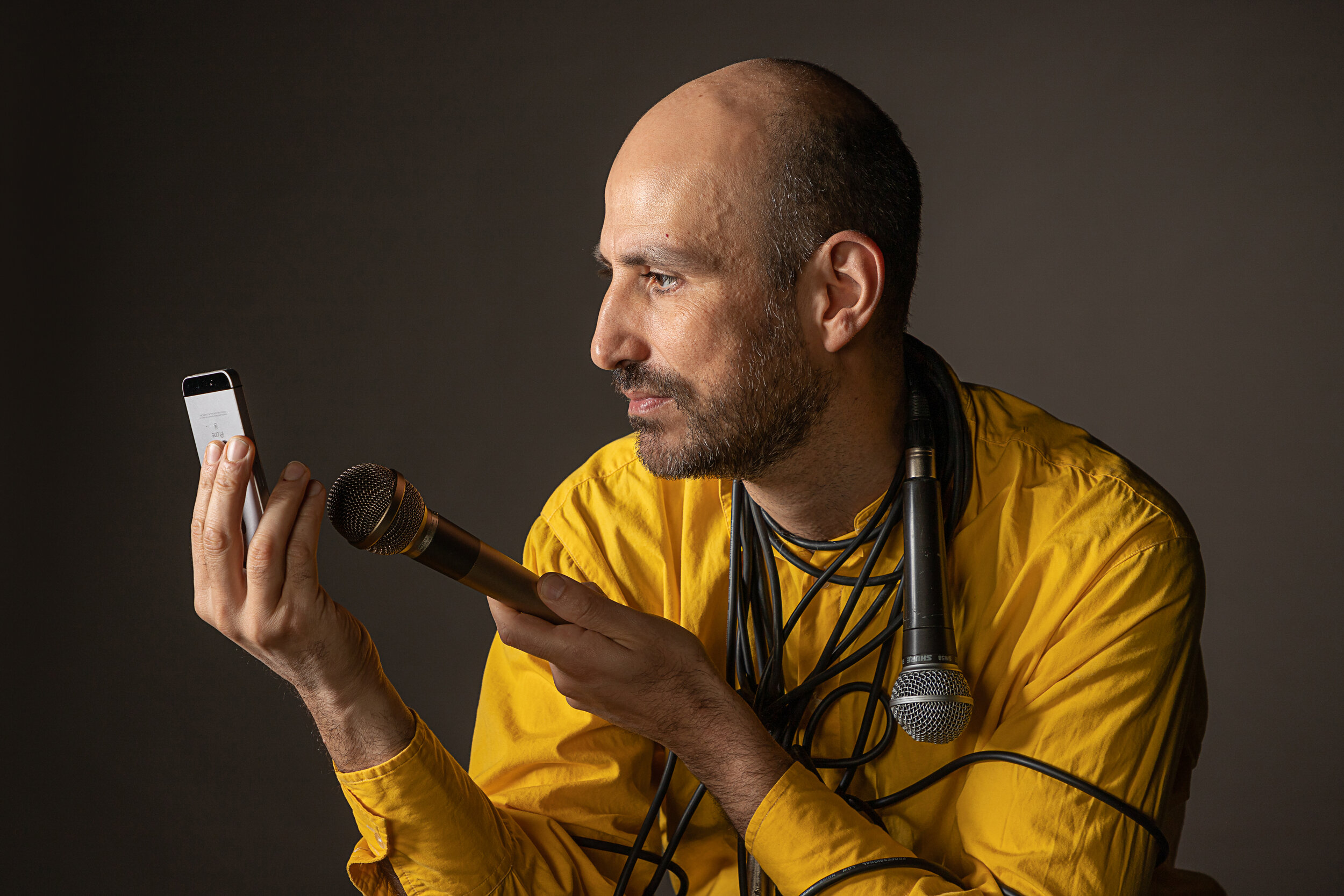 Man with a shaved head and beard wearing a yellow shirt, holding a microphone and looking at a smartphone, with multiple microphone cables around his neck, against a dark background. Jon Cohen. Jon Cohen Experimental.