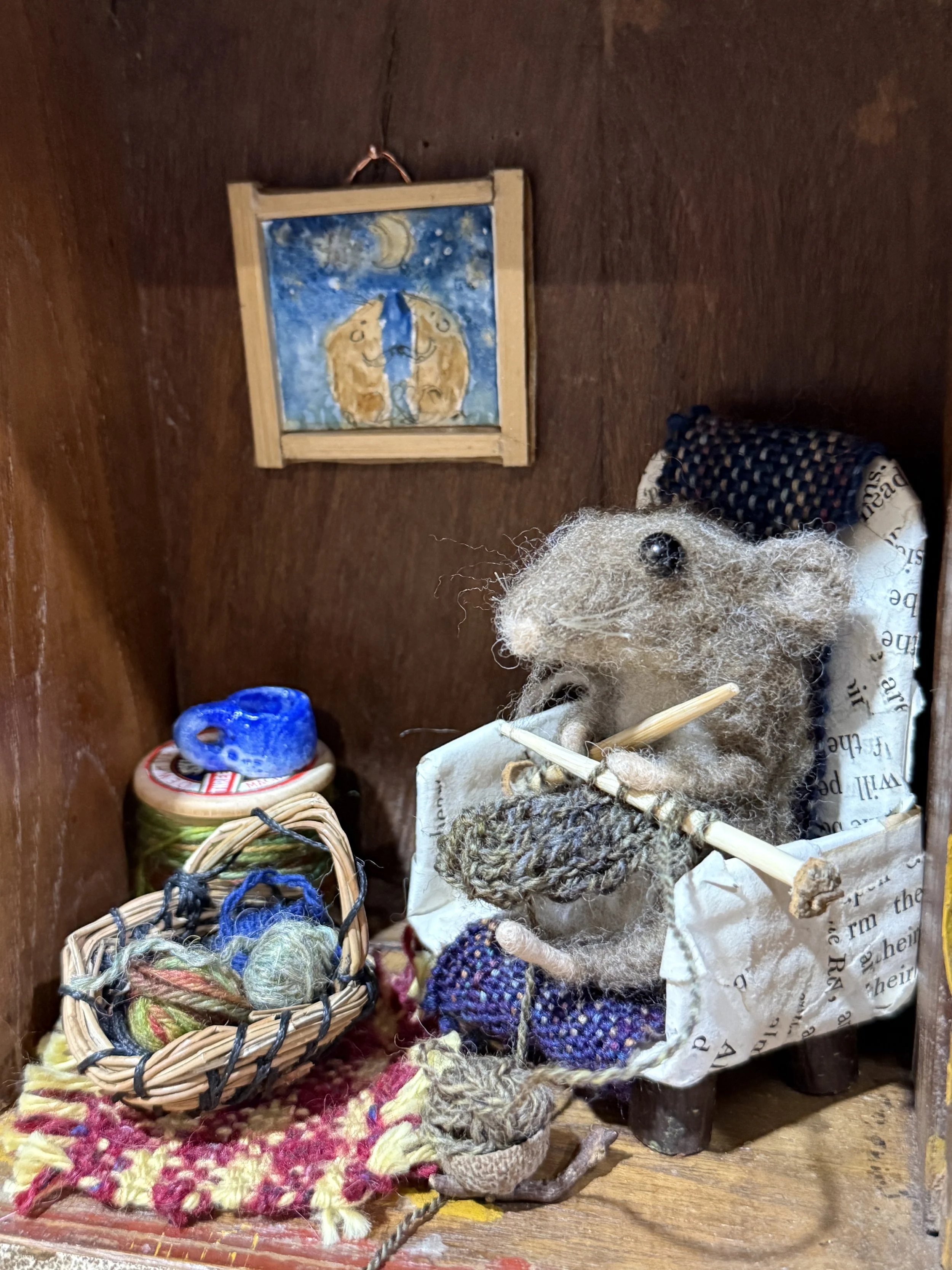 Vintage clock case of a mouse knitting in their chair. Handmade papier-mache  chair with wooden legs, tiny knitting, cotton reel table with a raku fired hand thrown mug. Handwoven pine needle basket with balls of wool, little painted picture on the w