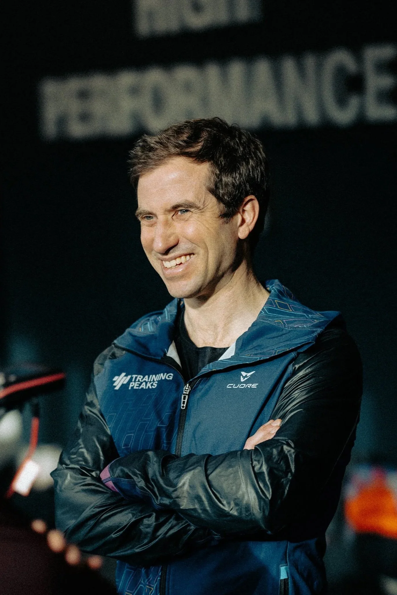A male trail runner in athletic gear running on a rocky mountain trail with snowy peaks in the background under a blue sky.