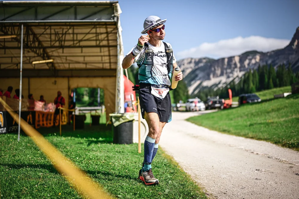 An ultrarunner leaving a Lavaredo Ultra Trail aid station with a refilled bottle in hand, Dolomite peaks behind