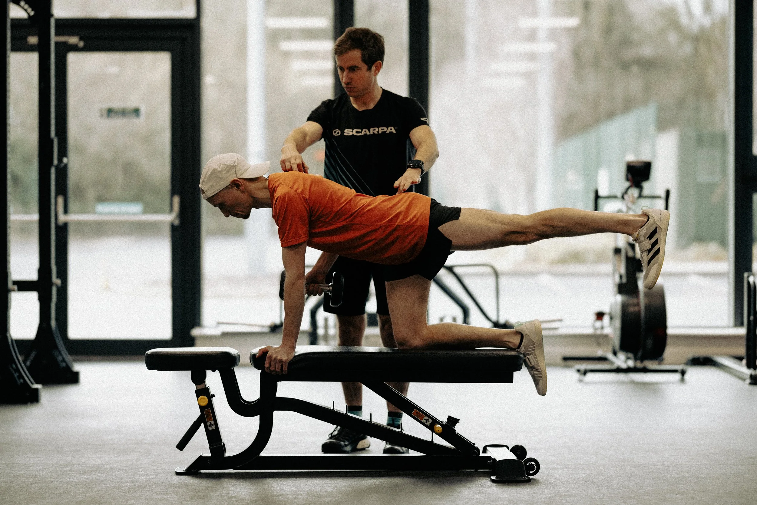 A woman preparing to exercise sitting on a bench in a gym with weights and workout equipment in the background.