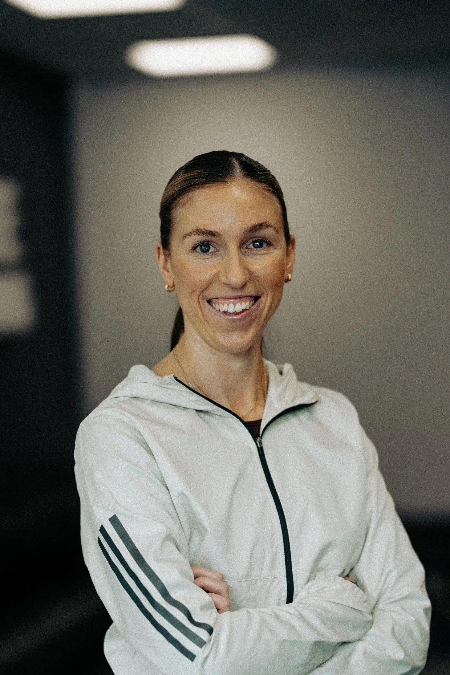 A woman in athletic wear stands with arms crossed in front of a dark wall with illuminated 3D white lettering that says 'ELITE TRAINING' and the woman is smiling.