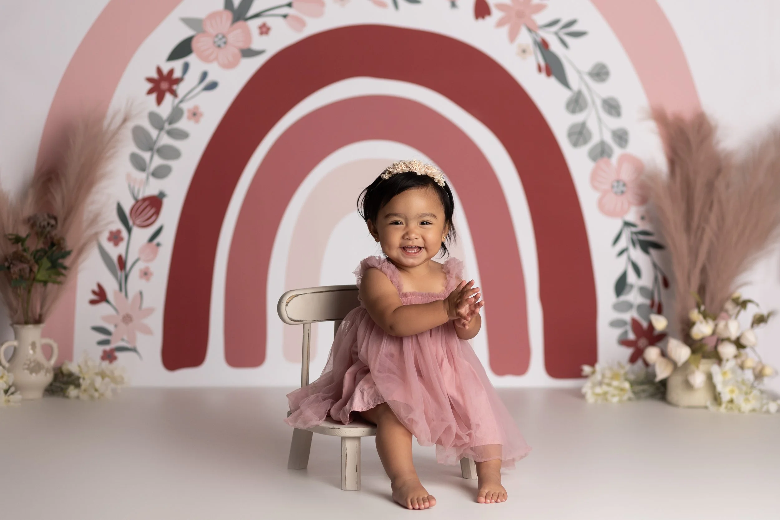 One year old girl in a pink dress smiling in front of a pink rainbow backdrop during a first birthday milestone session in Austin TX.
