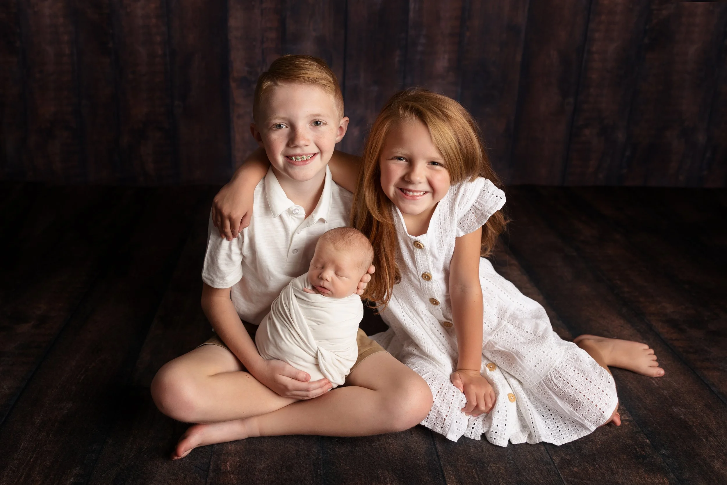 A heartwarming sibling portrait featuring two proud older siblings holding their newborn baby during a family newborn session. Sibling portraits are carefully guided to create safe, natural interactions and lasting family memories.