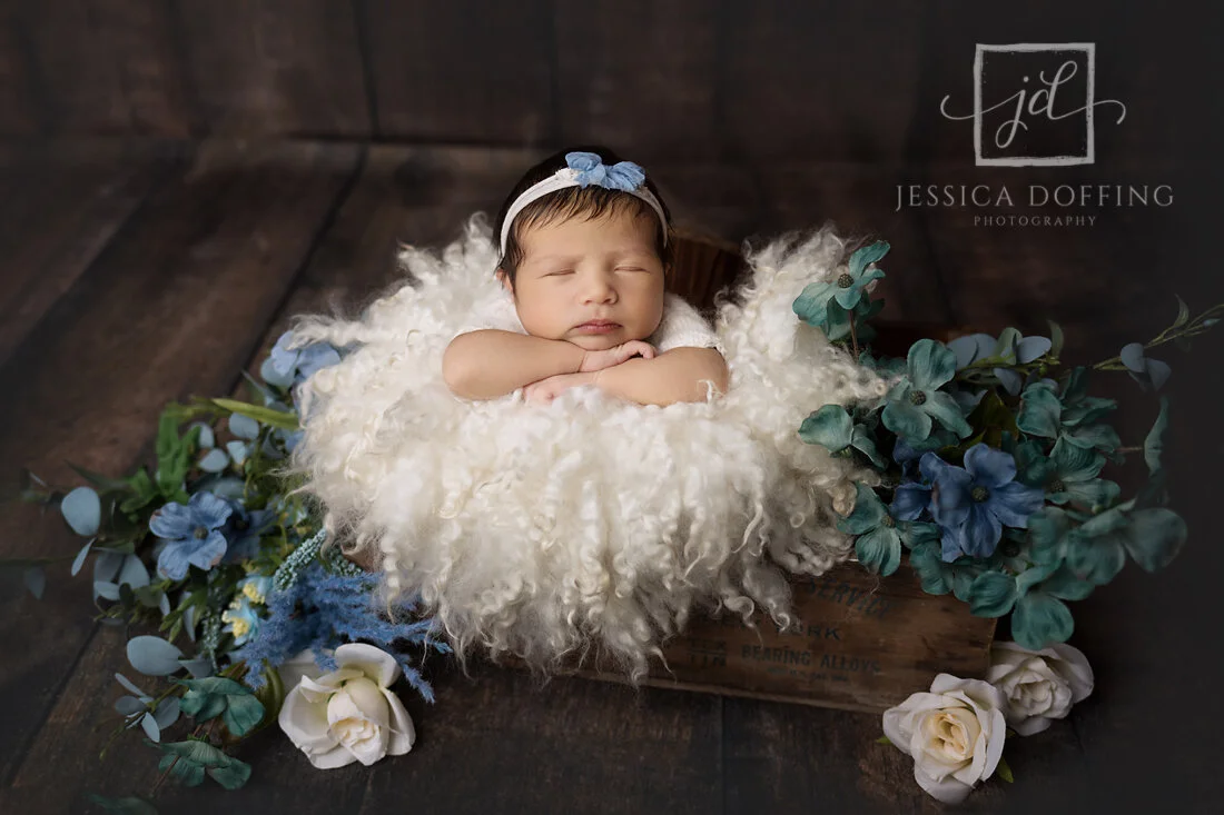 Newborn baby peacefully sleeping in a wooden crate surrounded by white flokati and blue floral accents during a professional newborn photography session in Austin, Texas.