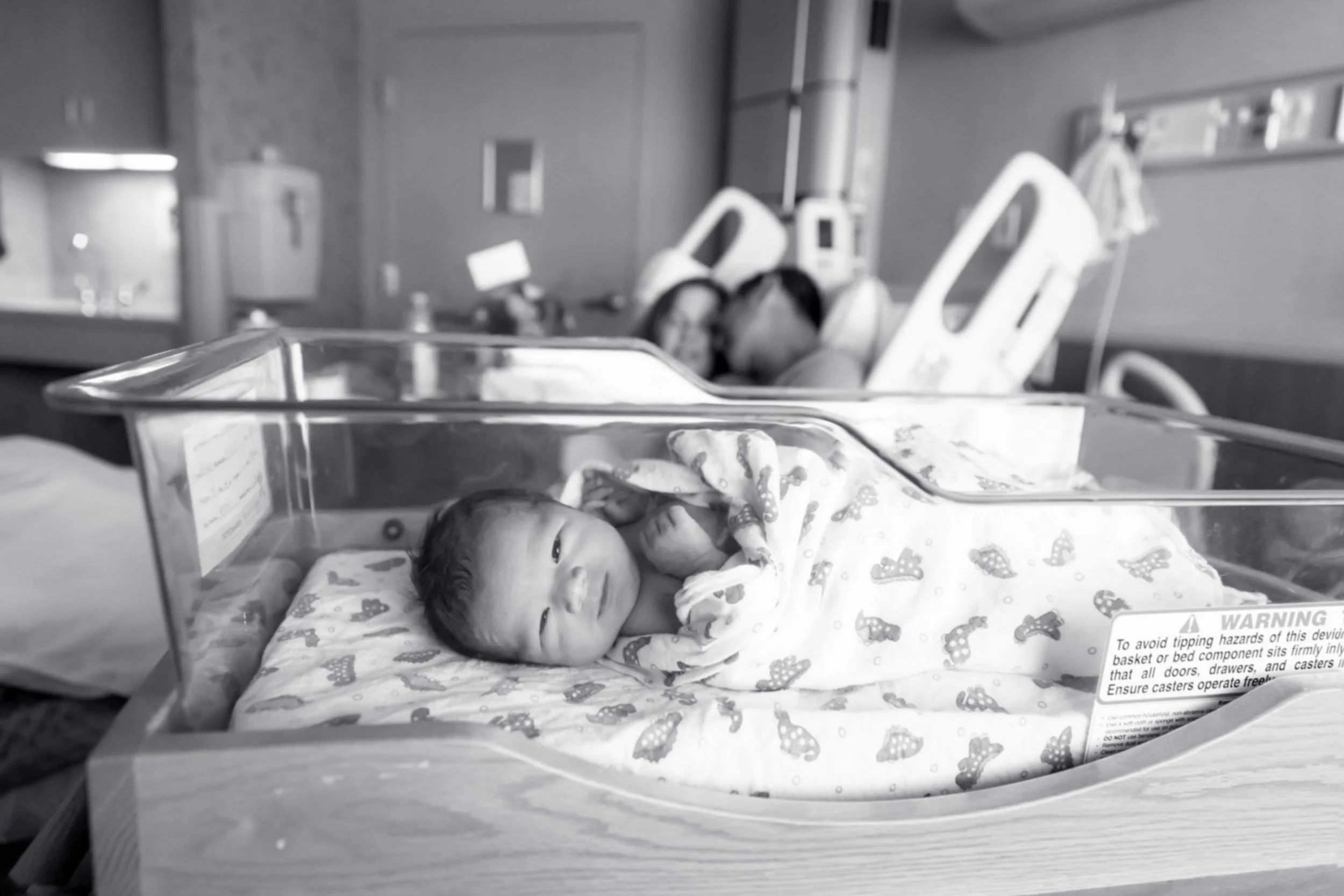Newborn baby in hospital bassinet with parent snuggling in the background