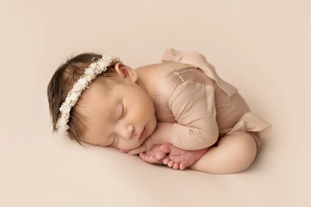 A peacefully sleeping newborn baby girl posed in womb/taco position on a soft neutral backdrop. Simple newborn photography highlights your baby's delicate features while creating timeless heirloom portraits.
