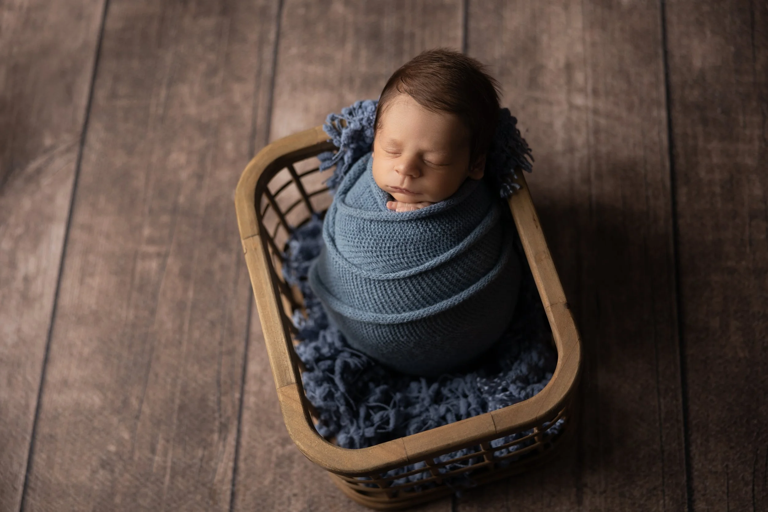 newborn baby boy wrapped in blue blanket sleeping in wooden basket on dark wood floor