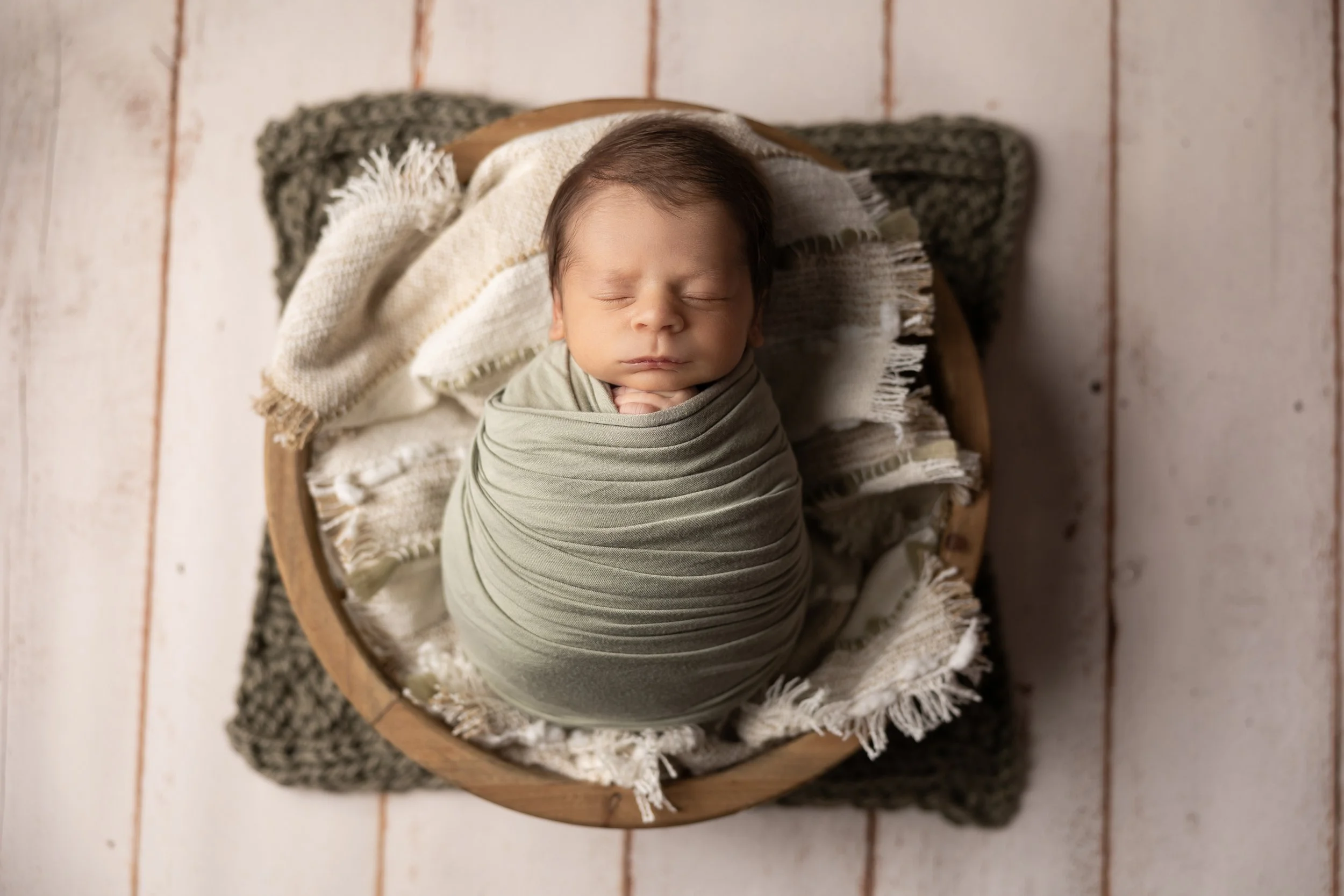 newborn baby boy wrapped in soft green fabric sleeping in wooden bowl with neutral textures