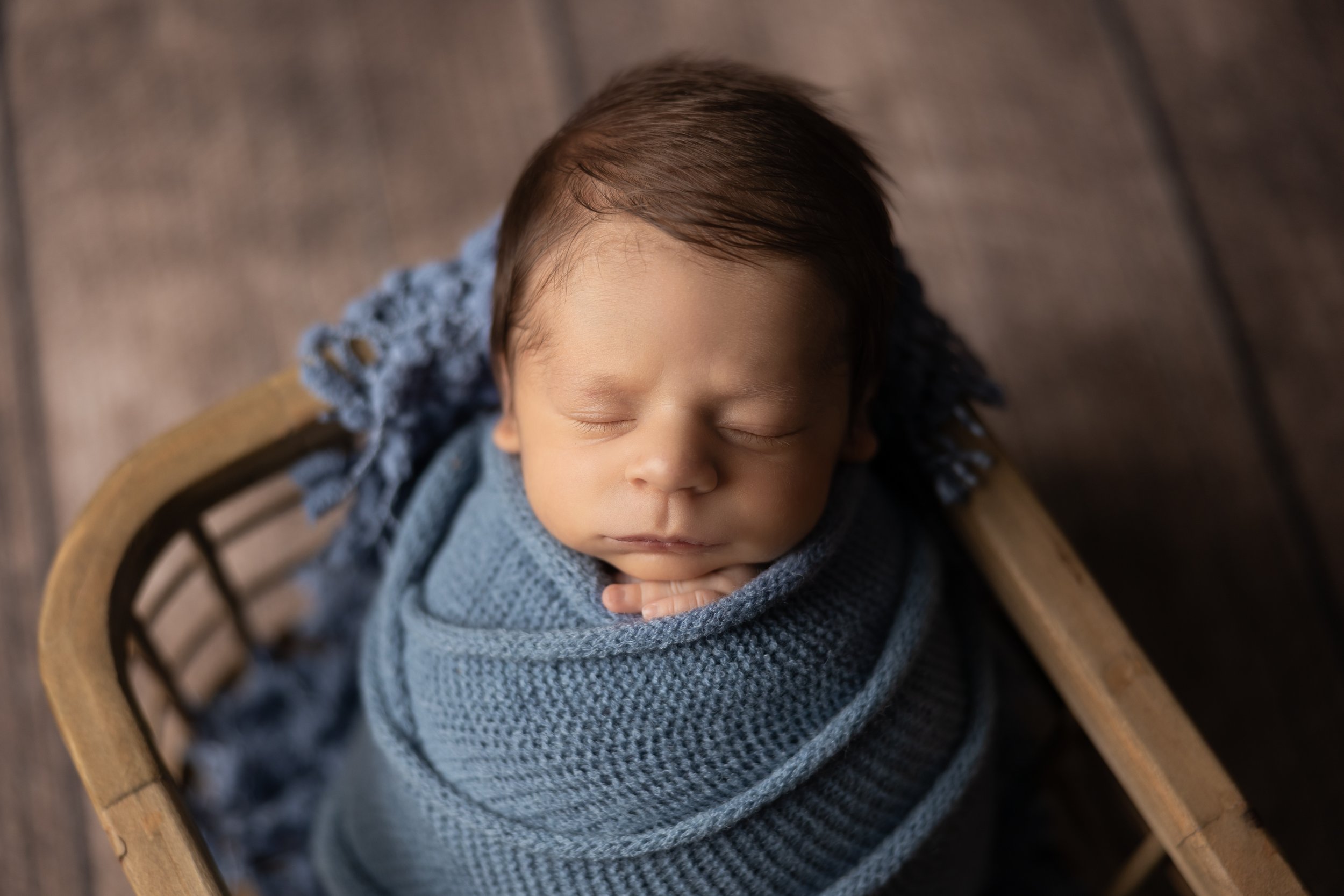 close-up of sleeping newborn boy wrapped in blue knit fabric resting in wooden prop