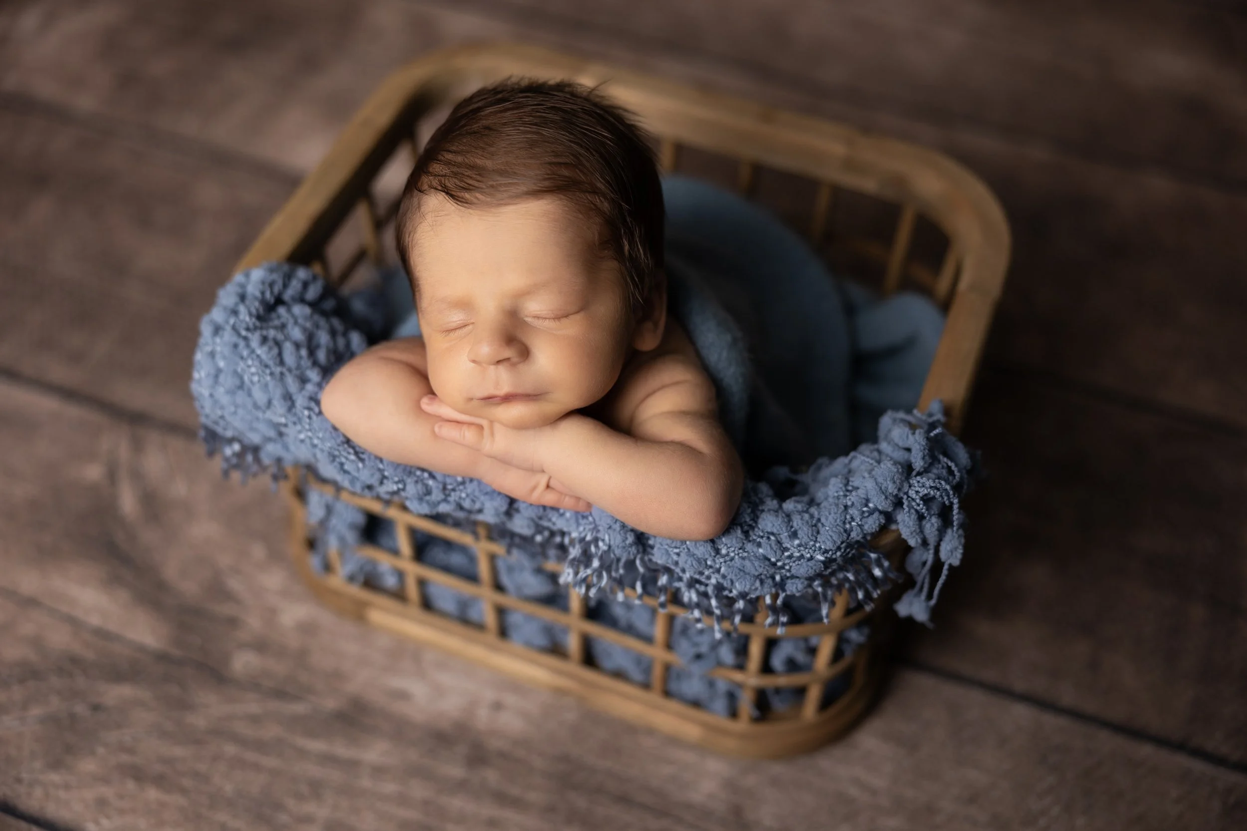 sleeping newborn boy resting chin on hands in basket with soft blue texture