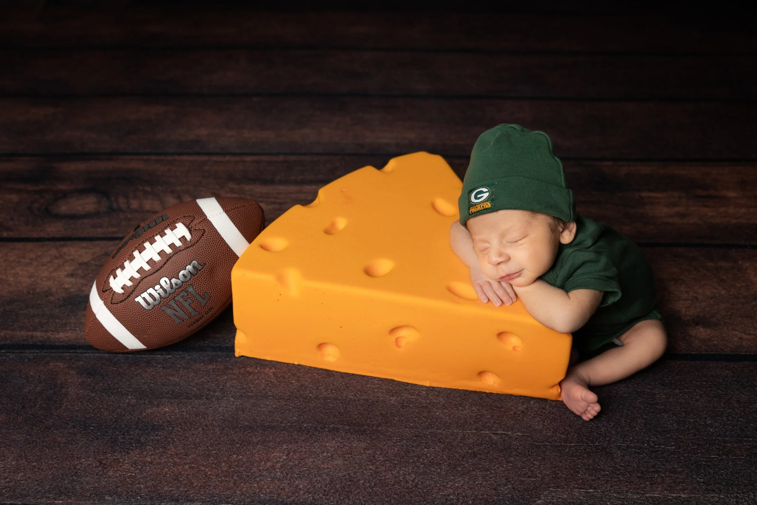 newborn baby dressed in green outfit posed next to football and cheese block prop